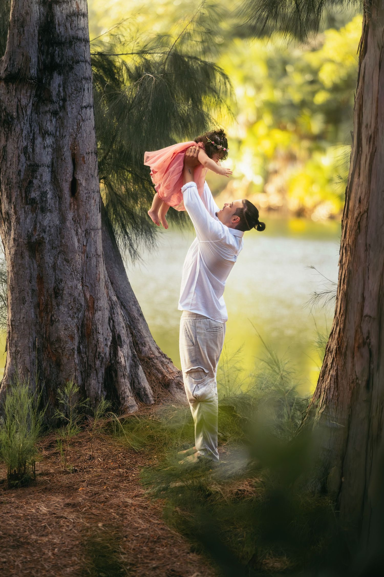 Father joyfully lifting young daughter in pink dress among trees by a sunlit lake in a lush park.