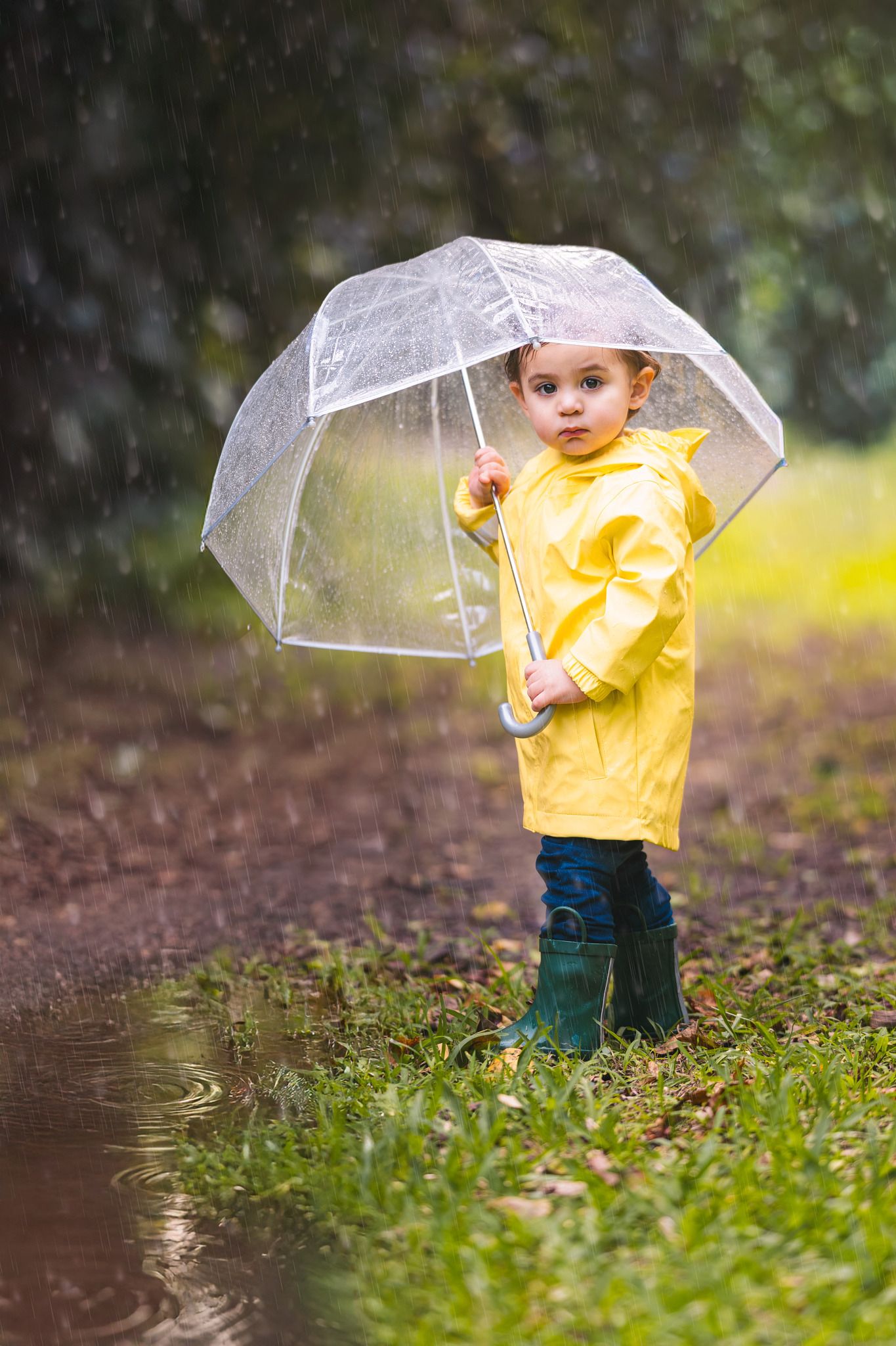 A small child in a bright yellow raincoat plays in the rain while holding a clear umbrella on wet grass.