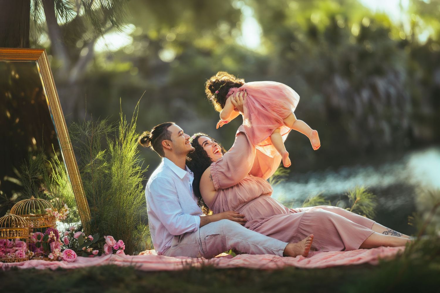 Happy family sitting outdoors on a blanket, parents lifting their young daughter in a pink dress near a serene garden.