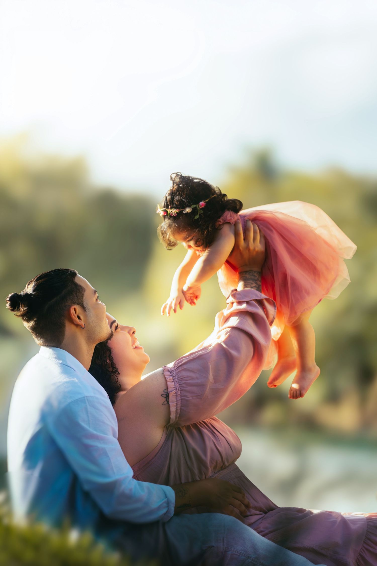 A happy family outdoors, parents lifting their young daughter in a pink tutu dress, bathed in warm golden sunlight.
