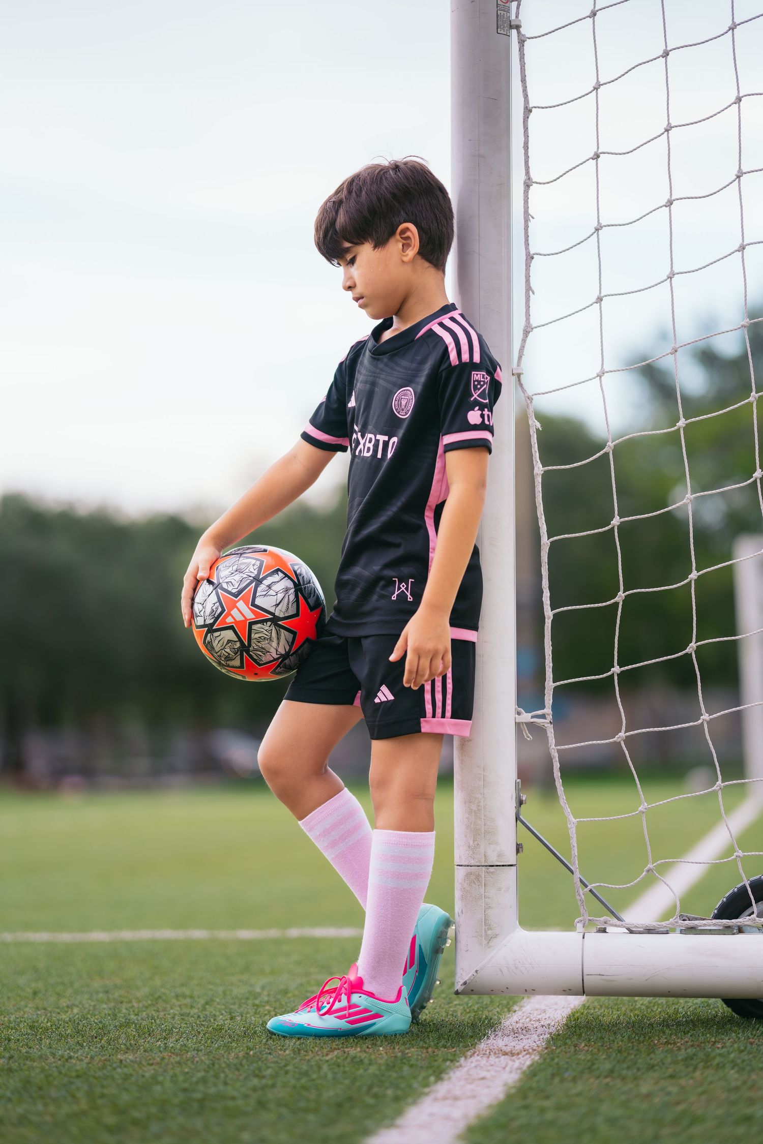 Young boy in black and pink Inter Miami soccer uniform holding a ball beside a goal post on a green field.