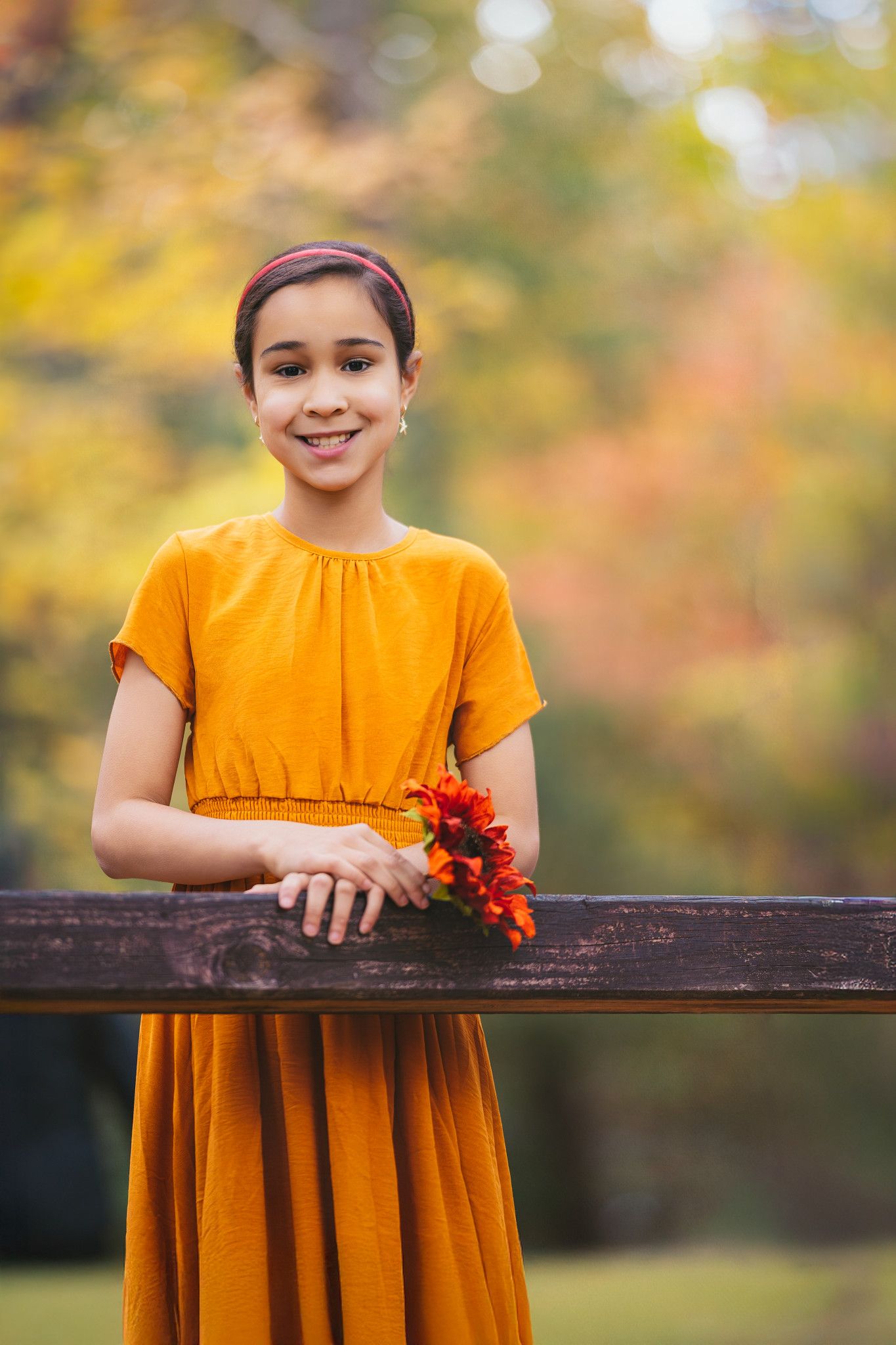 A smiling girl in an orange dress holds autumn flowers while leaning on a wooden fence in a fall park.