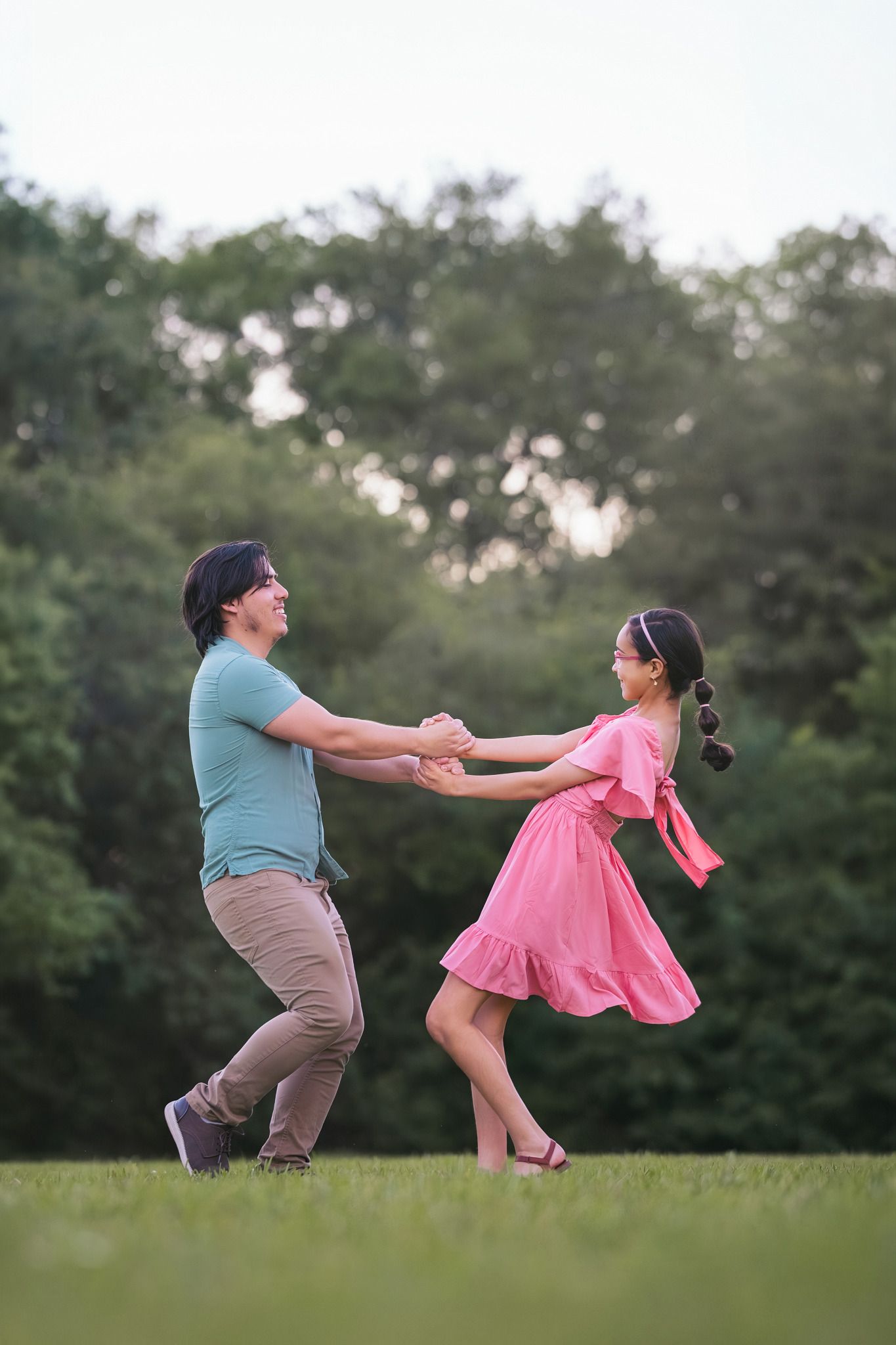 A joyful couple dances together on a green lawn, surrounded by lush trees, as the woman twirls in a pink dress.