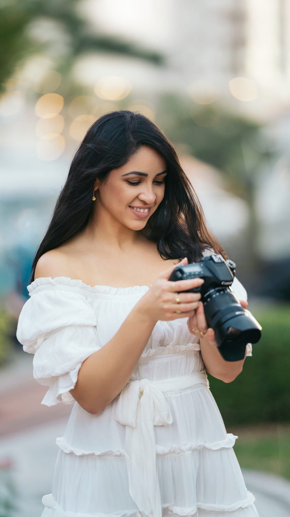 Woman in white dress reviewing photos on digital camera in a garden setting.