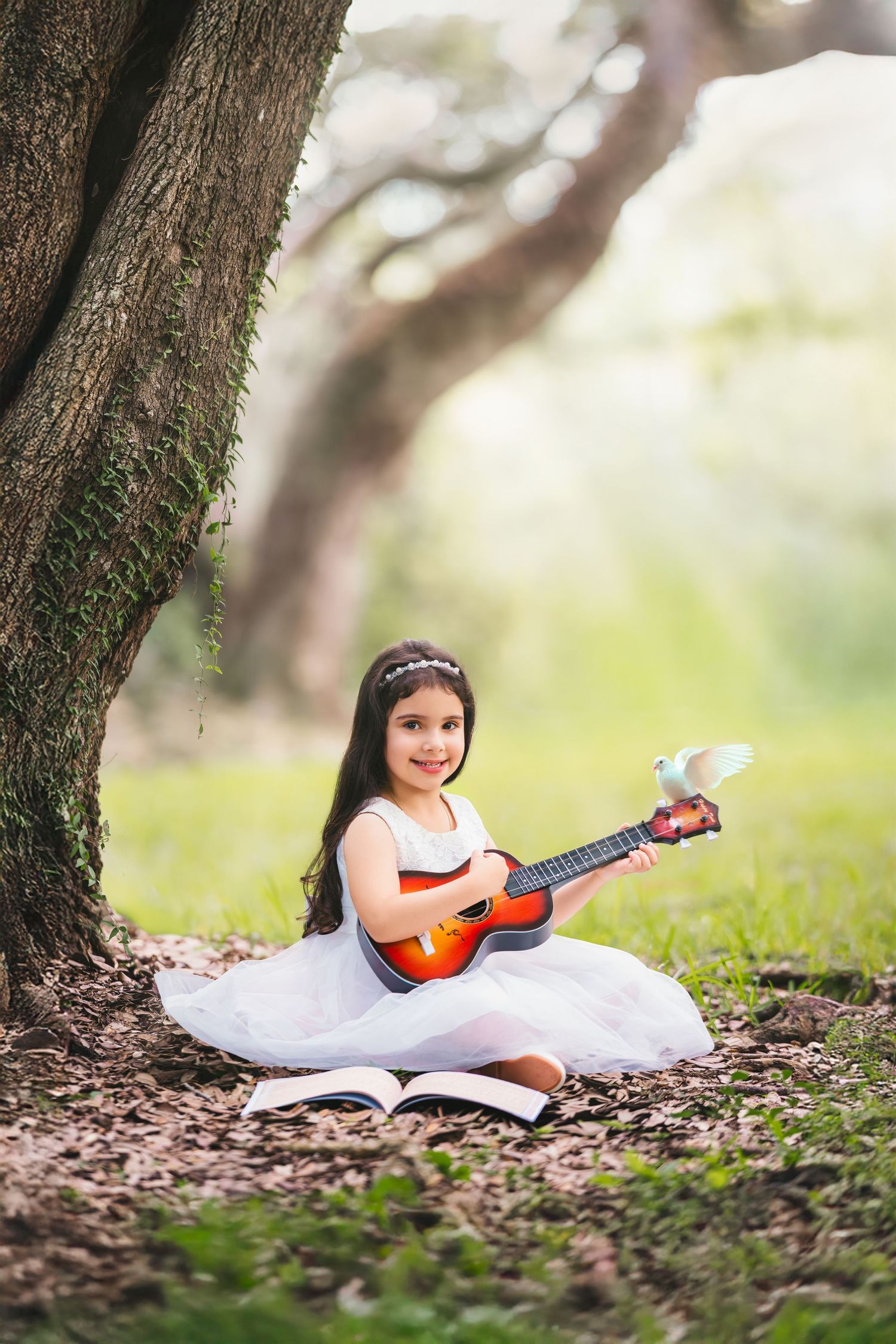 Young girl in white dress playing ukulele under a tree with a white dove perched on the instrument.