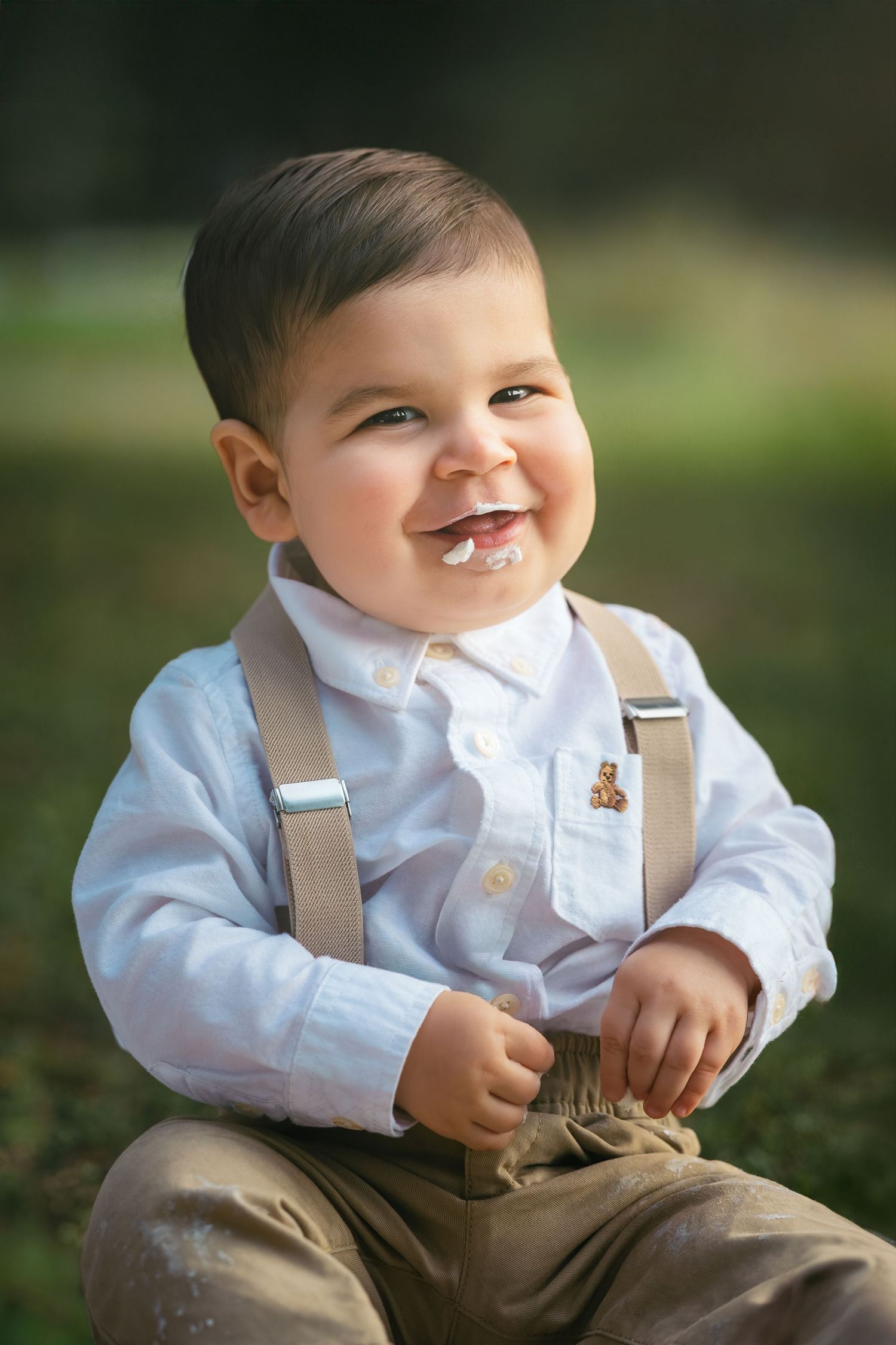 A smiling baby boy in white shirt with suspenders and a teddy bear pin sits outdoors with frosting on his face.