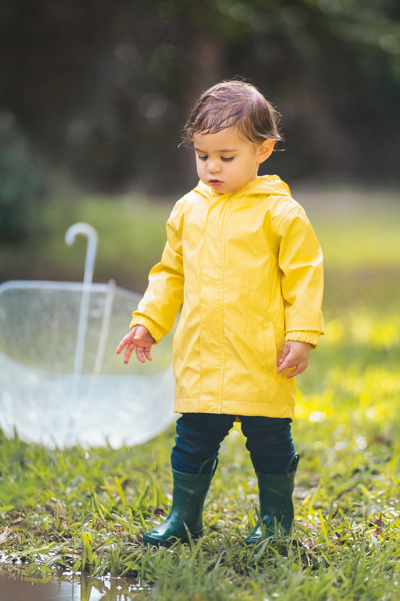 Toddler in yellow raincoat and green boots stands in puddle beside clear umbrella in lush green park.