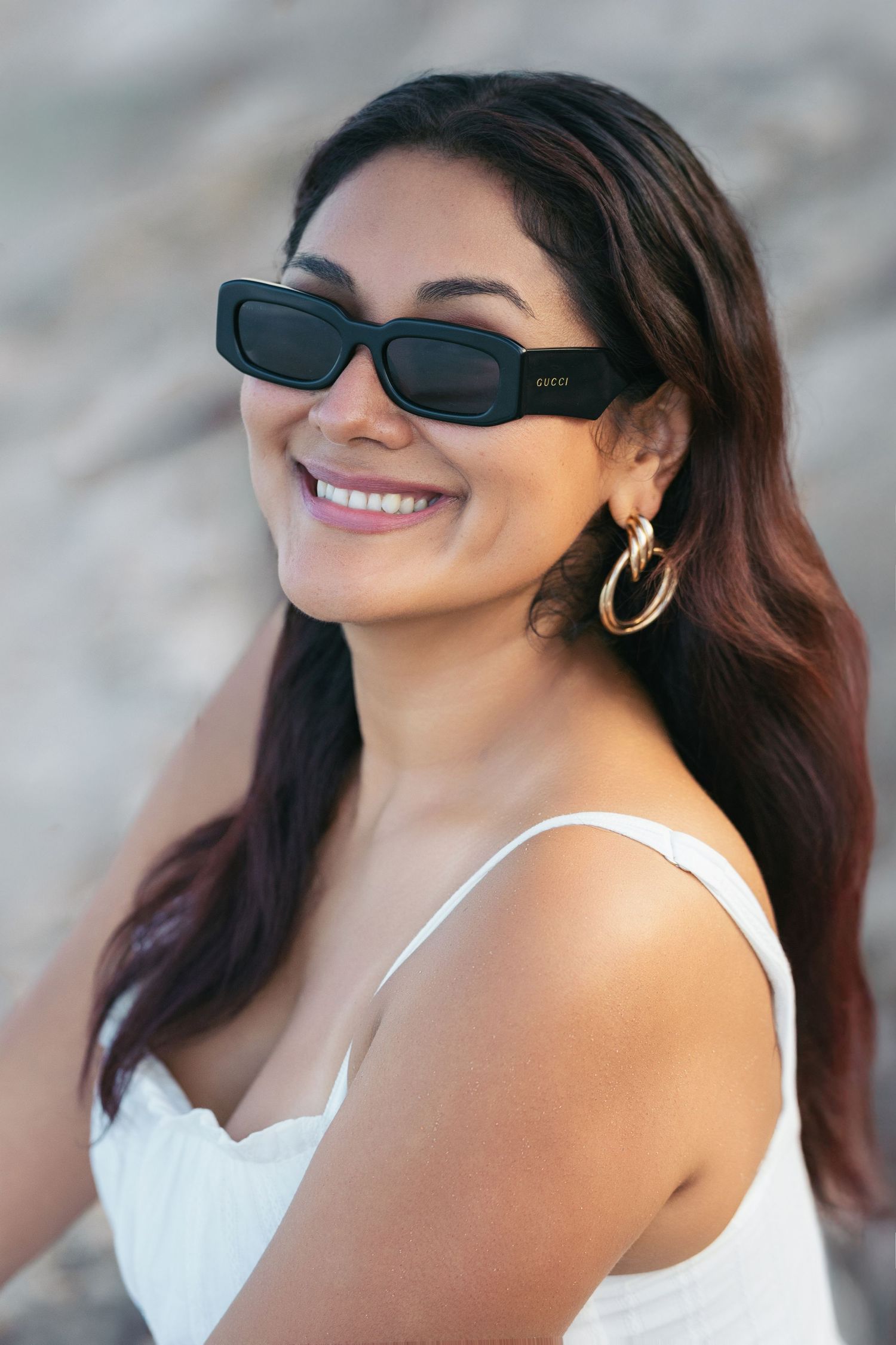 Smiling woman wearing black Gucci sunglasses and gold hoop earrings in a white top outdoors.