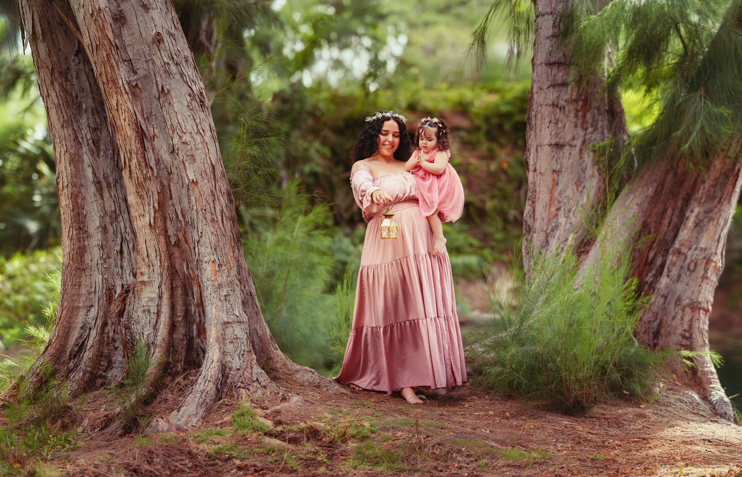 Mother and daughter in matching pink dresses with floral crowns posing between trees in a lush garden.