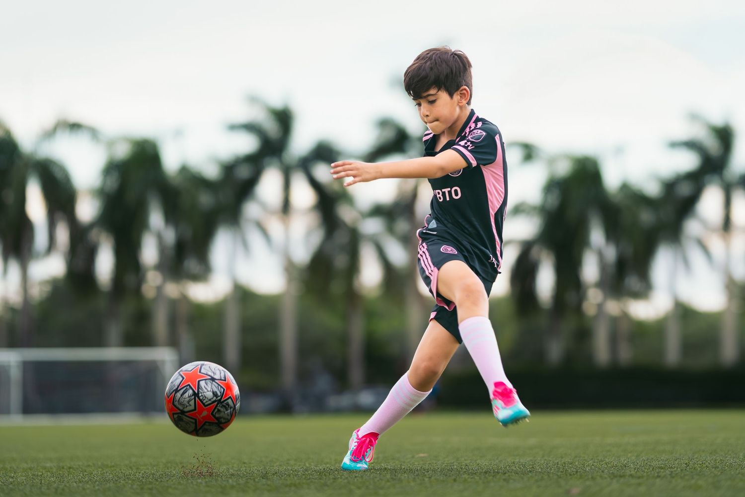 Young boy in pink and black soccer uniform kicking a red ball on a green field with palm trees in background.