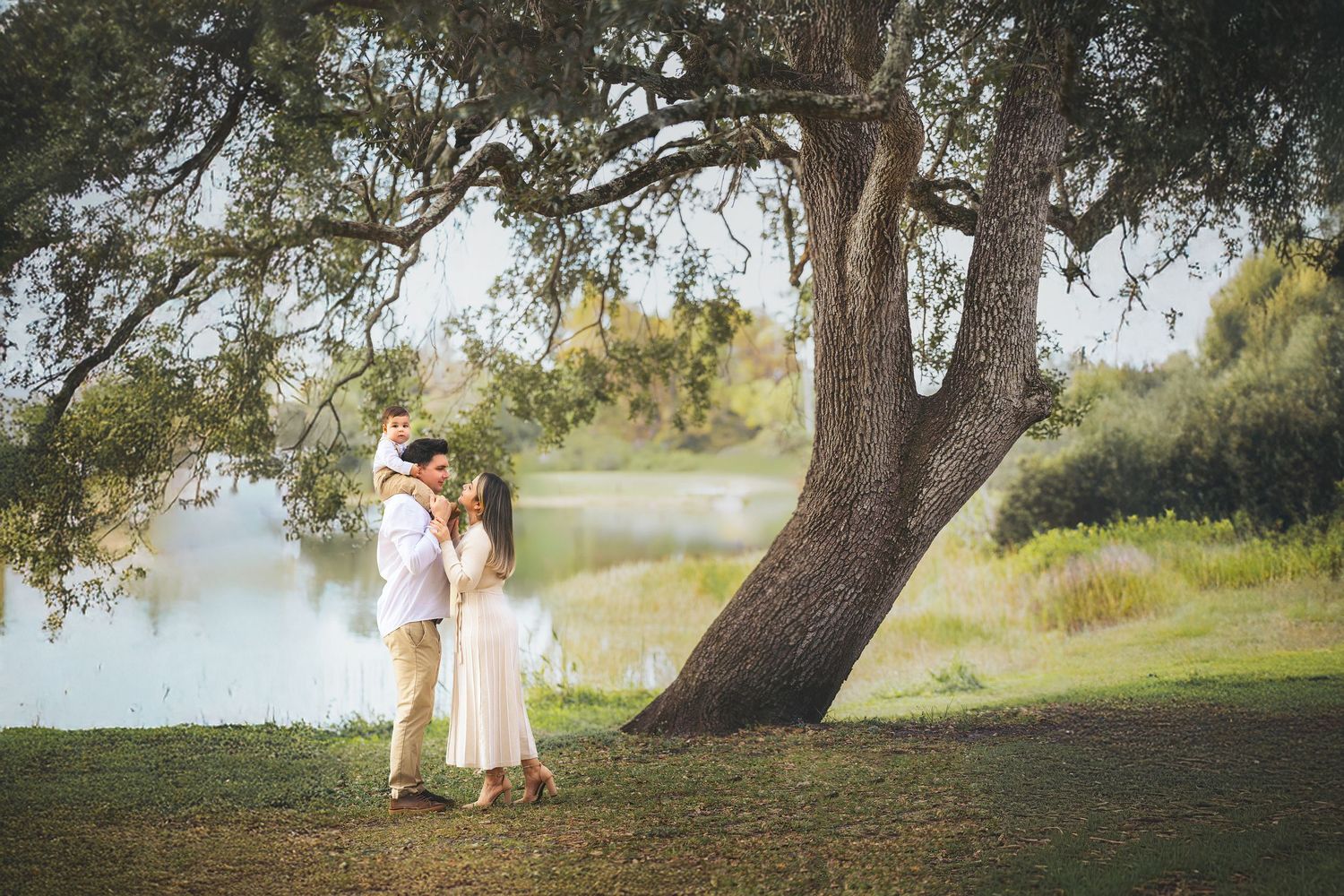 Couple embracing under a large oak tree by a serene lake in a lush green park setting.