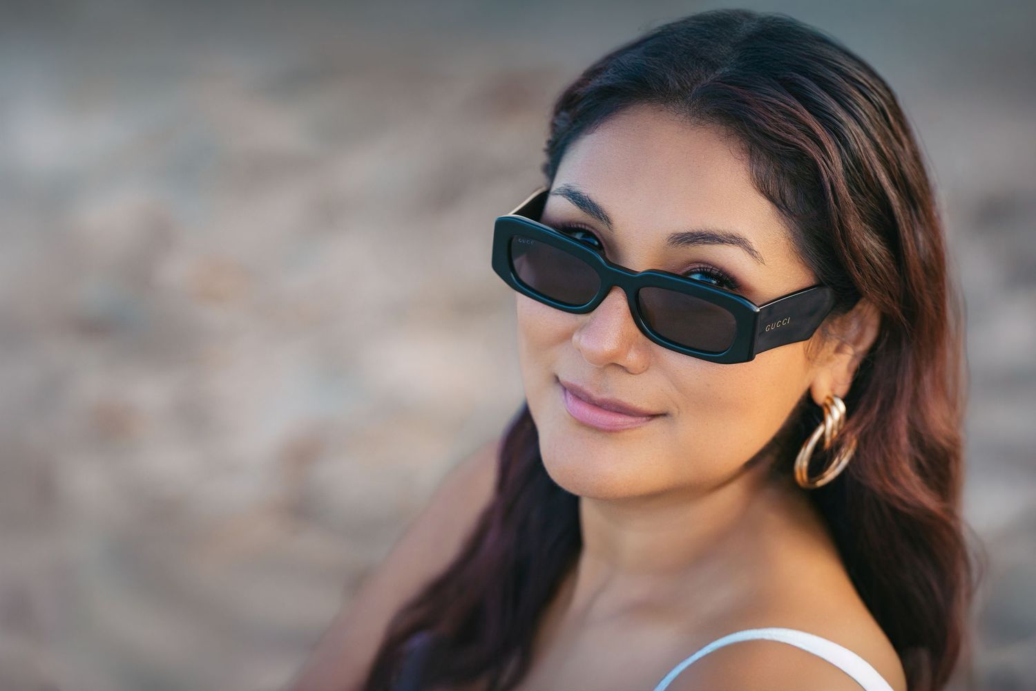 A person wearing trendy rectangular sunglasses and gold hoop earrings smiles while posing outdoors.