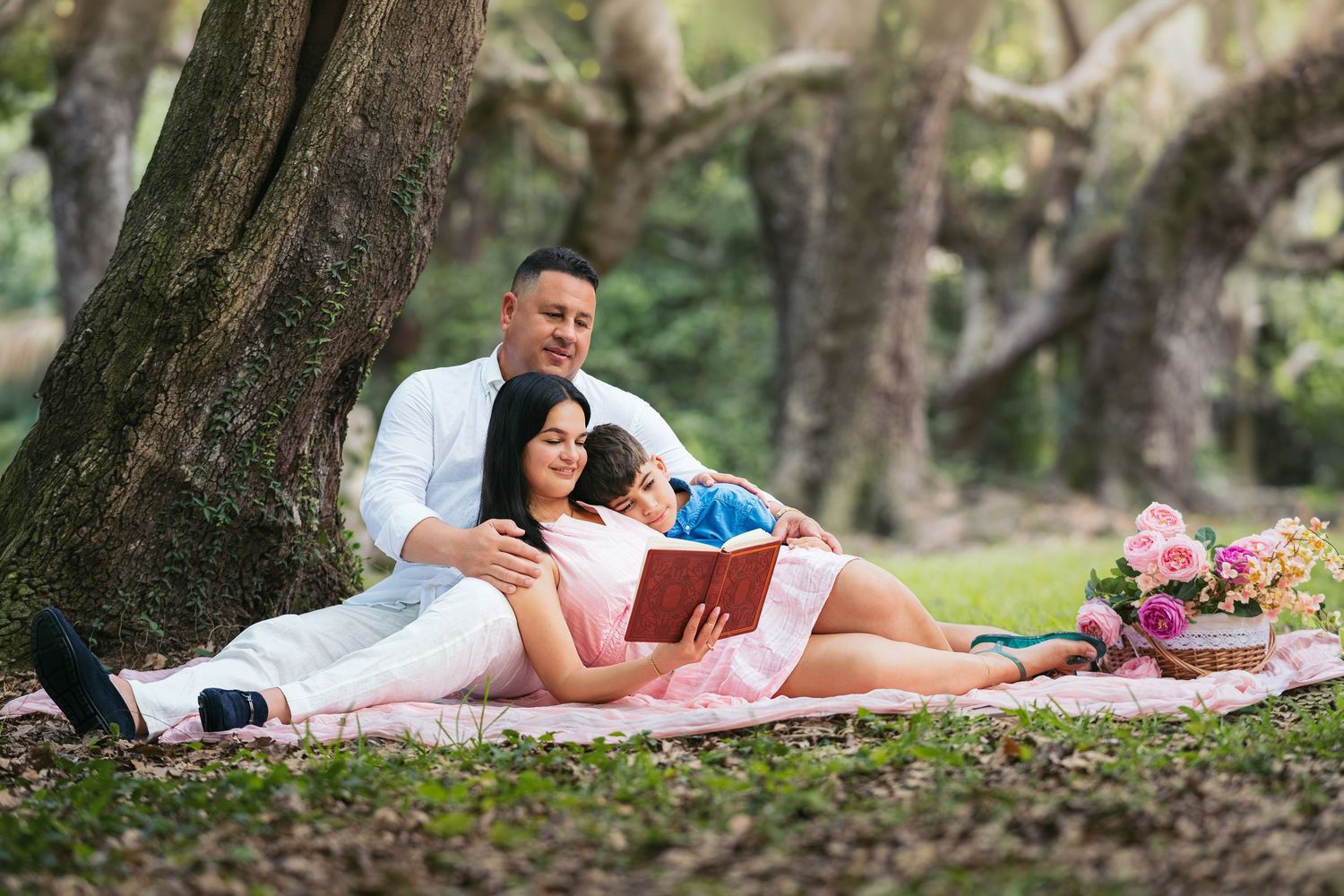 Family relaxing on a picnic blanket under trees, reading a book together in a lush green park.