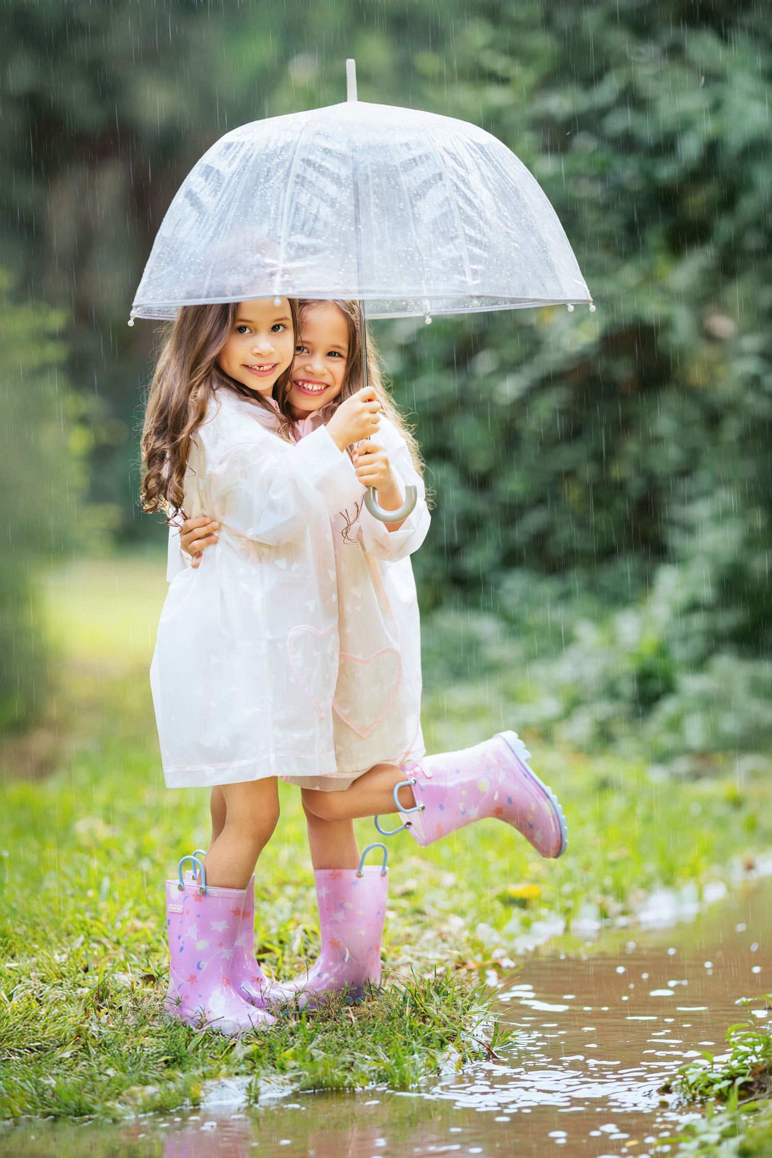 Two children in white dresses share a clear umbrella while wearing pink rain boots and standing in a puddle.