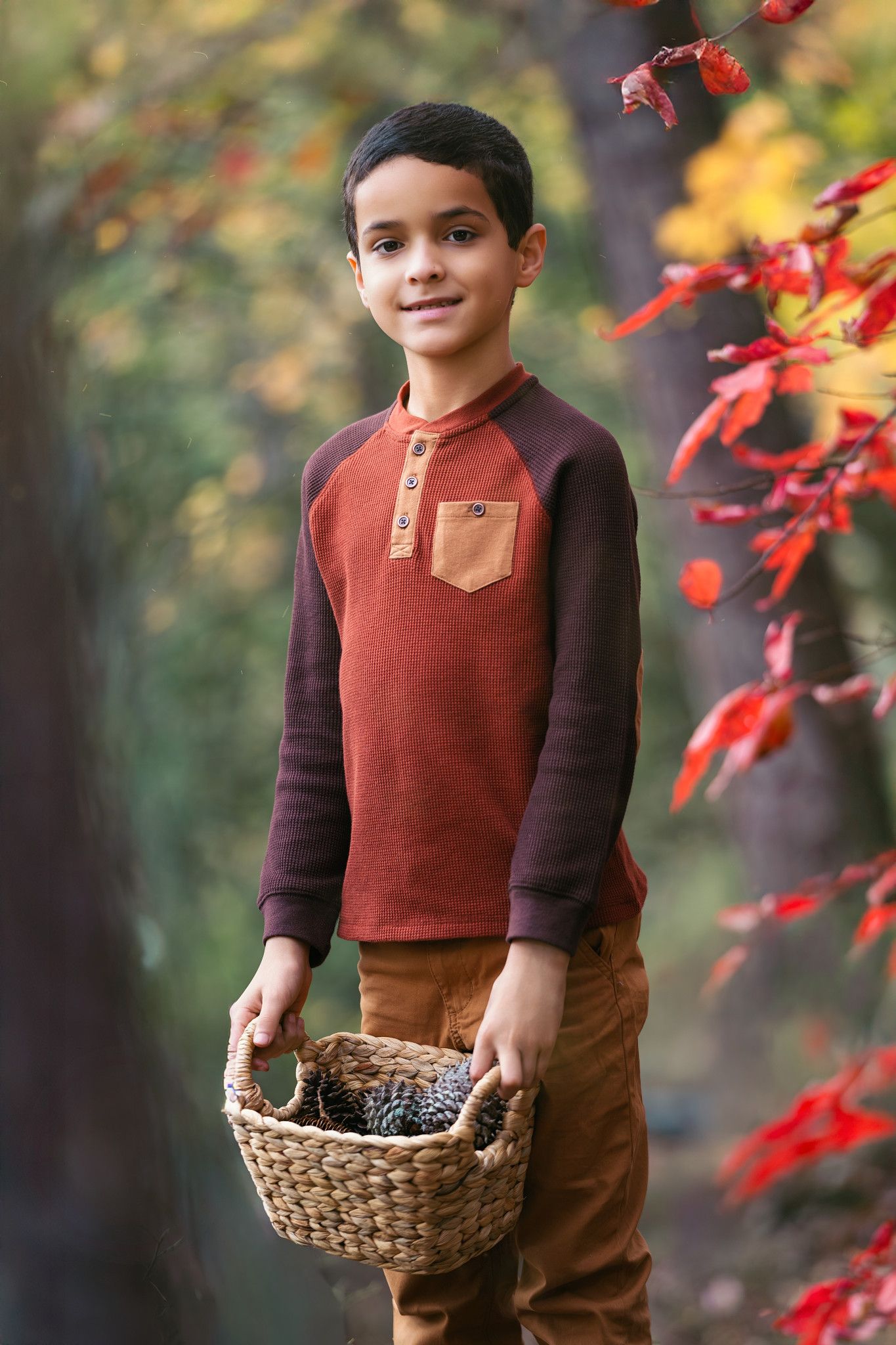 Boy in autumn forest holding wicker basket with pine cones, wearing brown raglan shirt and tan pants.