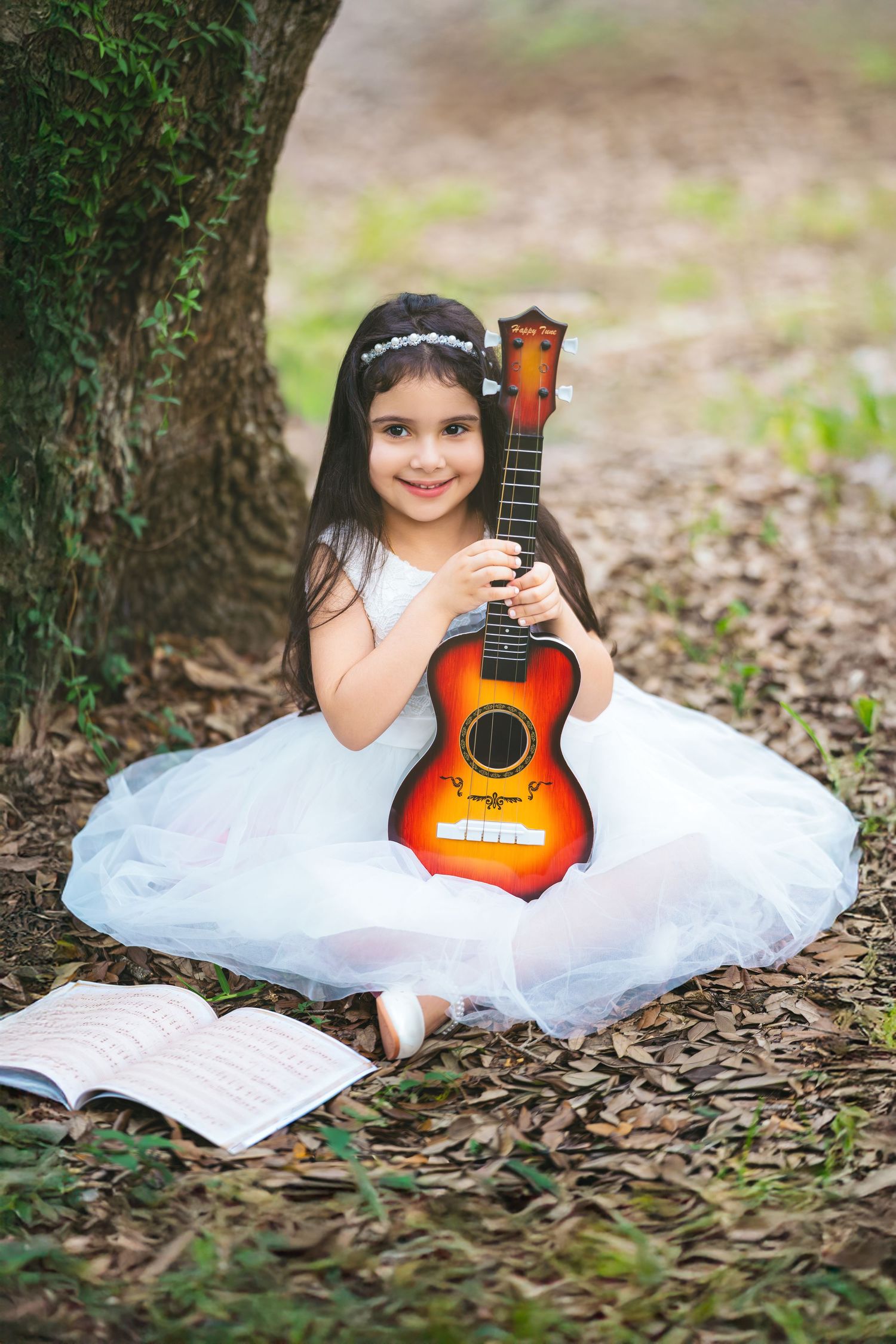 Young girl in white dress holding a colorful ukulele while sitting under a tree with sheet music nearby.