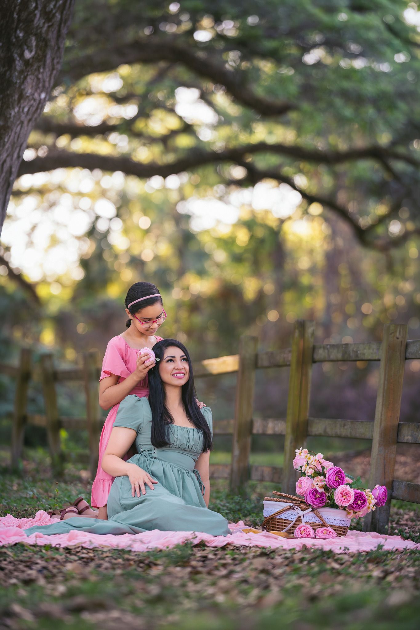 A mother and daughter share a tender moment on a pink blanket outdoors, surrounded by roses and lush greenery.