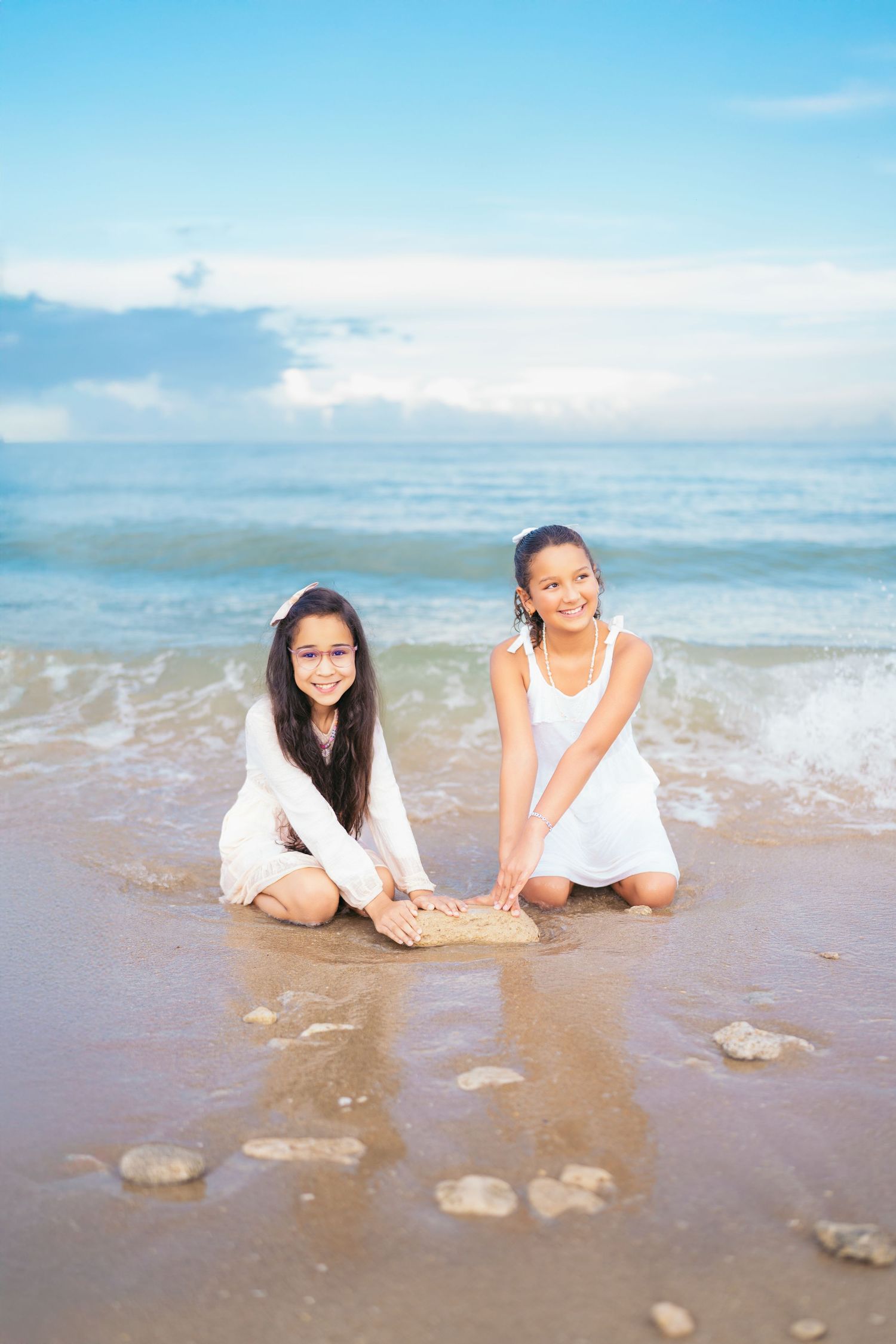 Two people in white outfits sit on wet beach sand with waves nearby.