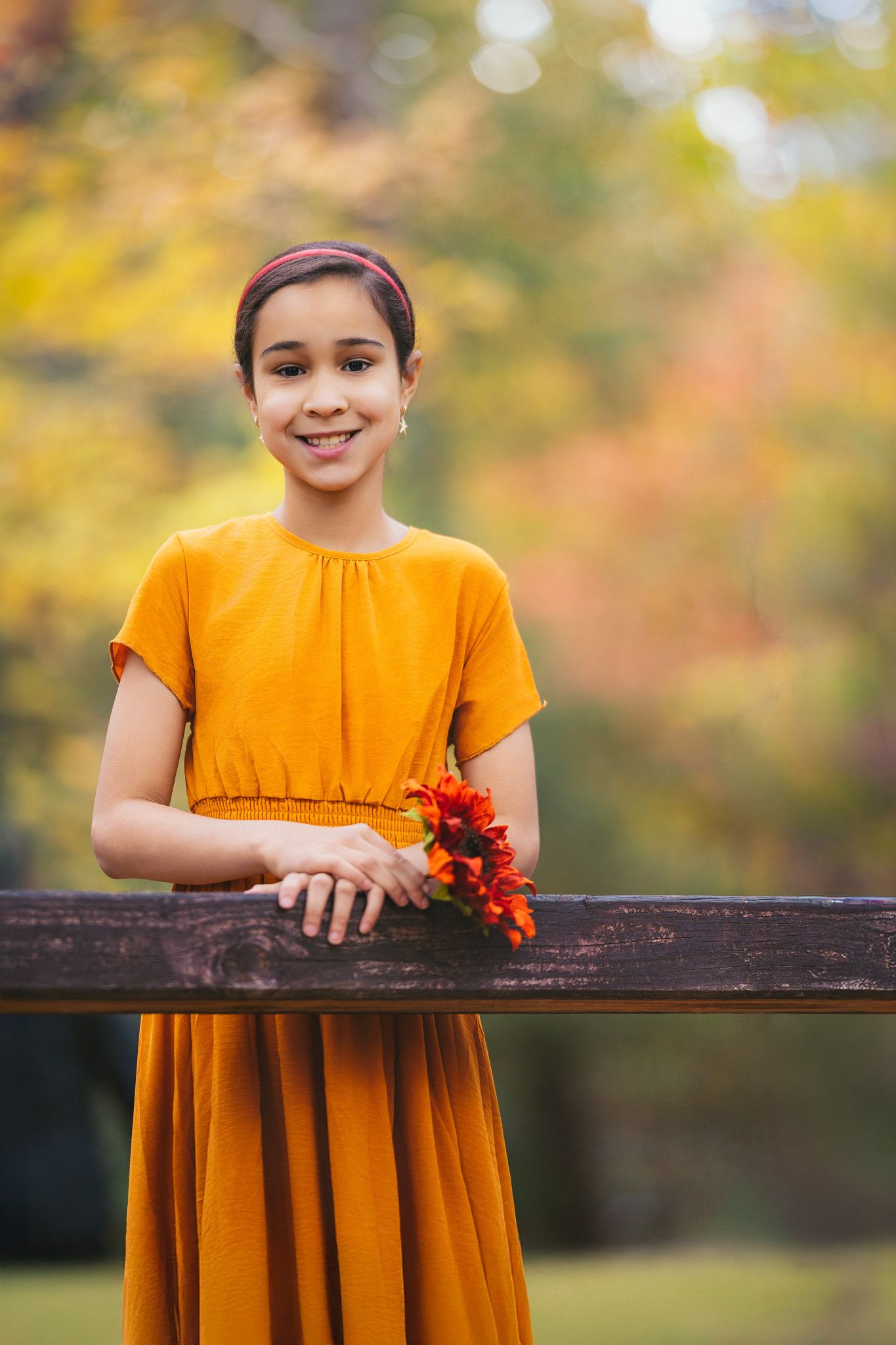 A child in a bright orange dress holds red flowers while leaning on a wooden railing in an autumn setting.