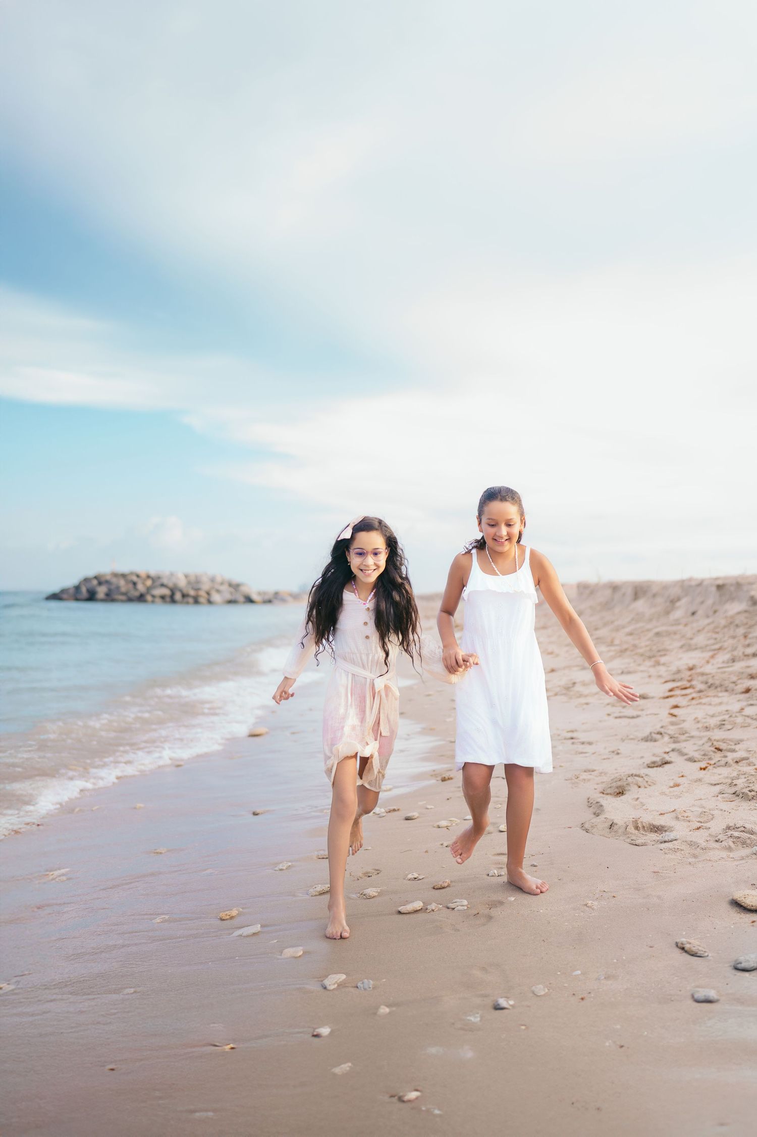 Two young girls in white dresses running barefoot on a sandy beach with ocean waves and rocky shore in background.