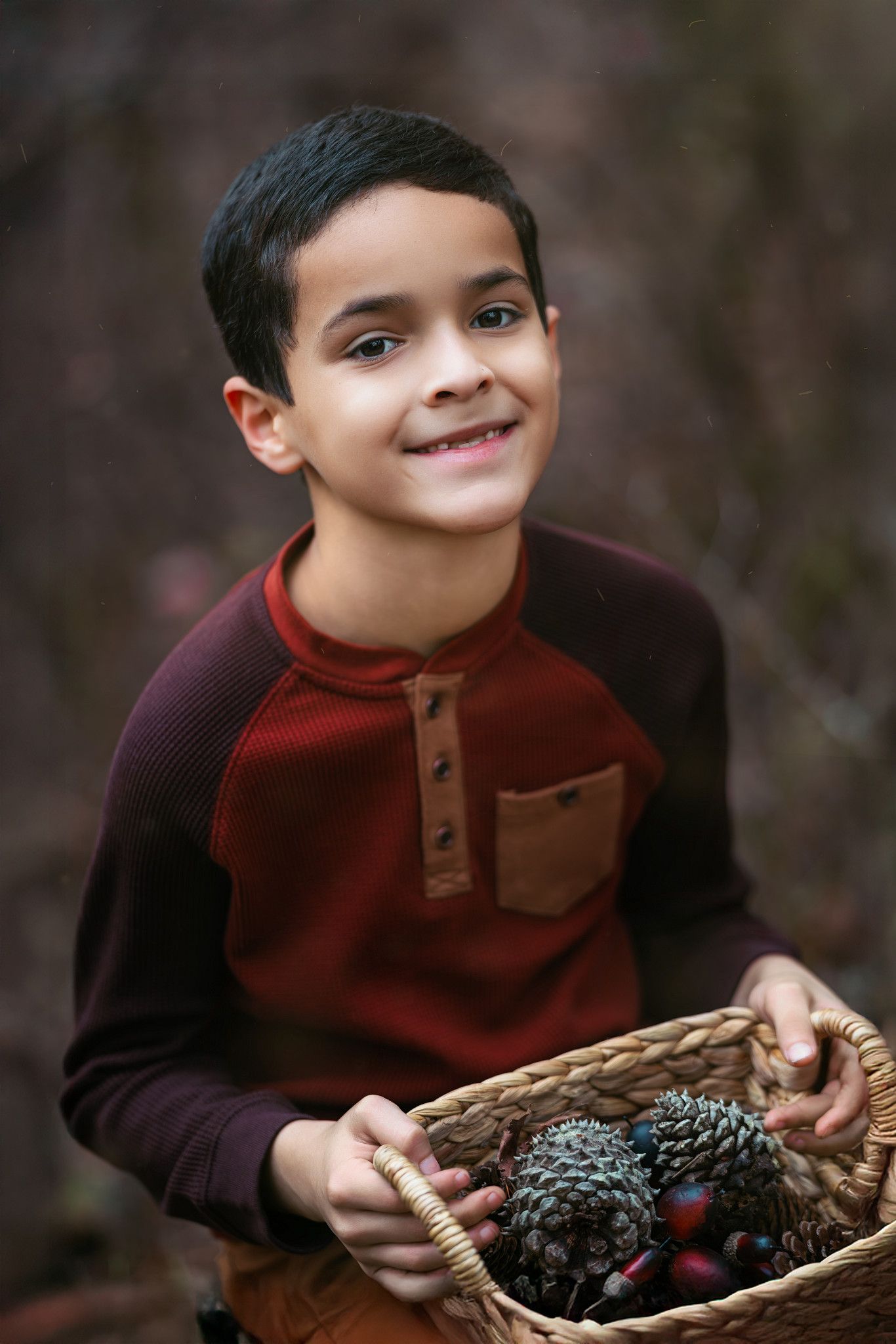Smiling young boy holding a wicker basket with pine cones outdoors in autumn wearing a red raglan shirt.