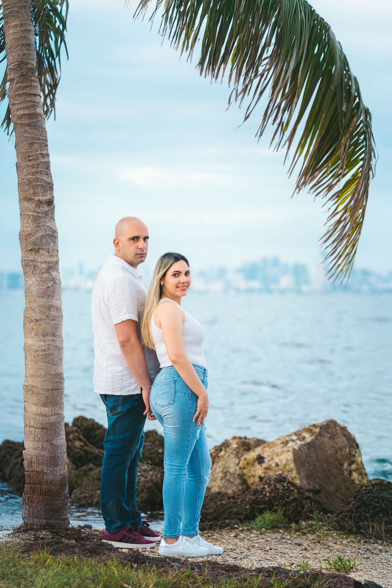 Couple standing back-to-back under a palm tree by the rocky waterfront with a city skyline in the background.
