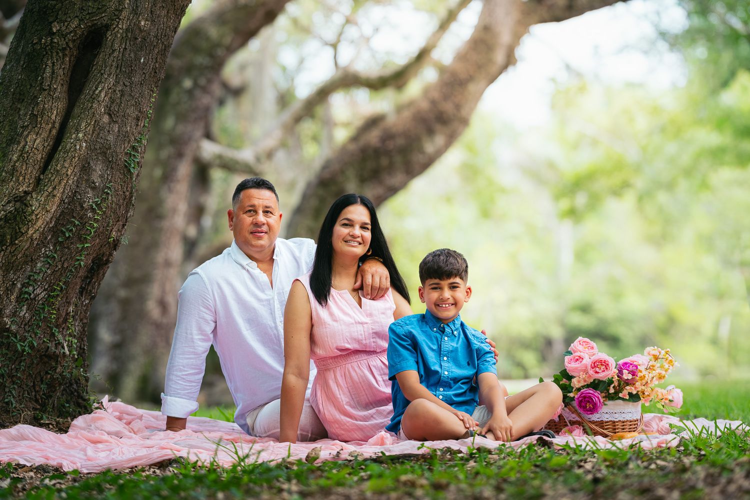 Family of four posing for a picnic photo under a large tree on a pink blanket in a lush green park.