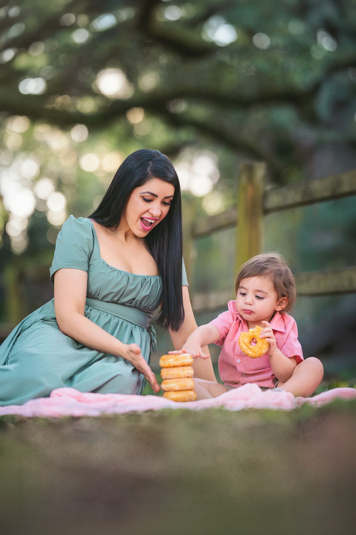 A woman in a teal dress sits outdoors with a toddler eating donuts on a pink blanket in a park.