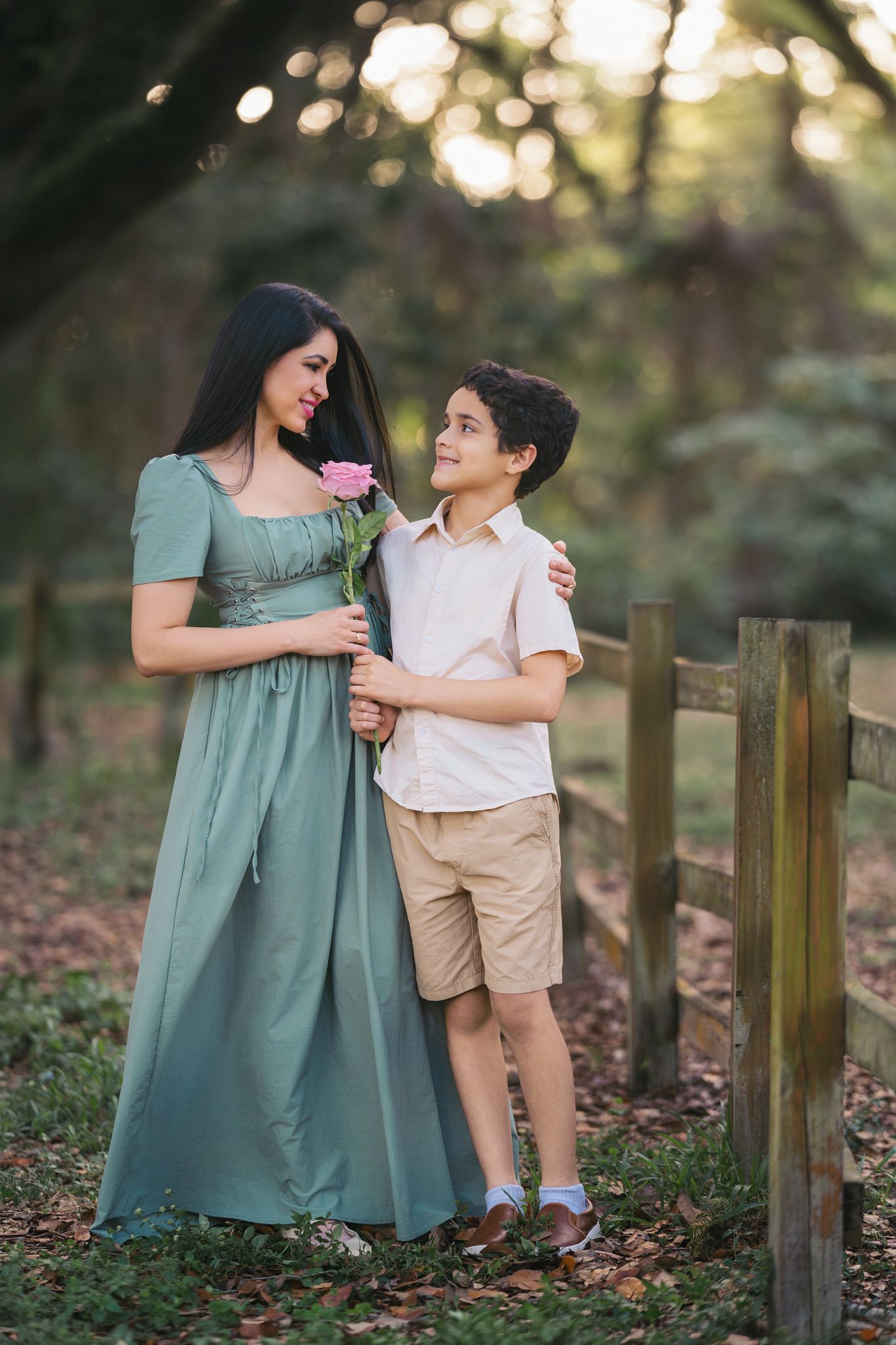 A woman in a green dress and a young boy holding a pink rose stand together near a wooden fence outdoors.