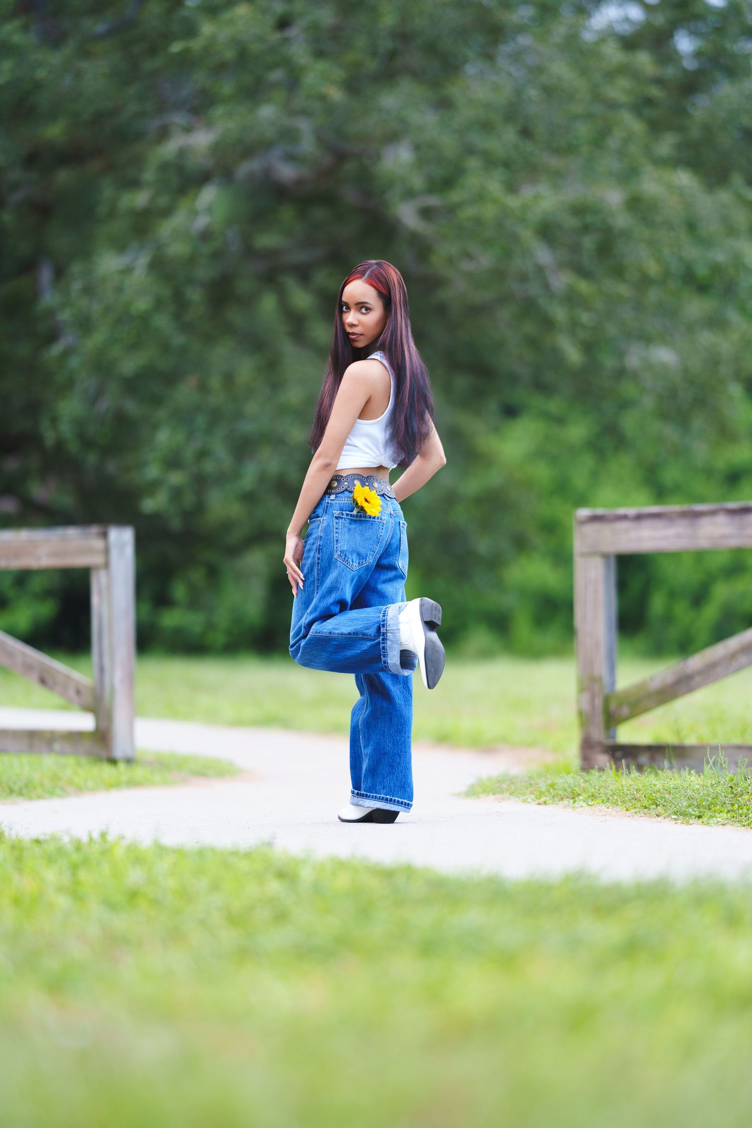 Young woman in wide-leg jeans and white crop top poses on a park path with wooden fences and green trees.