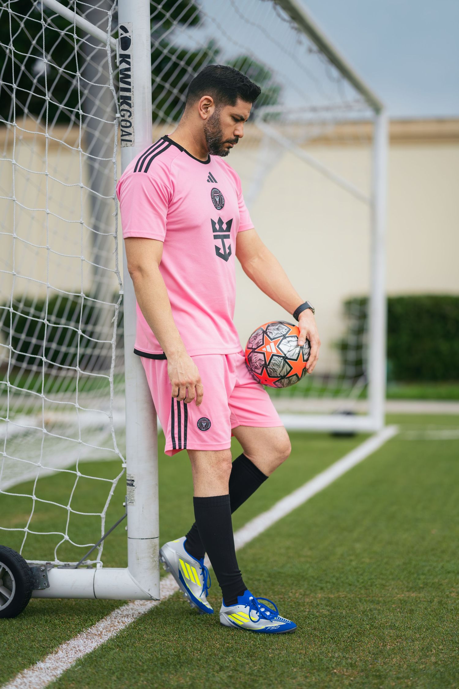 Soccer player in pink Inter Miami kit leaning against goal post holding a ball on a green field.