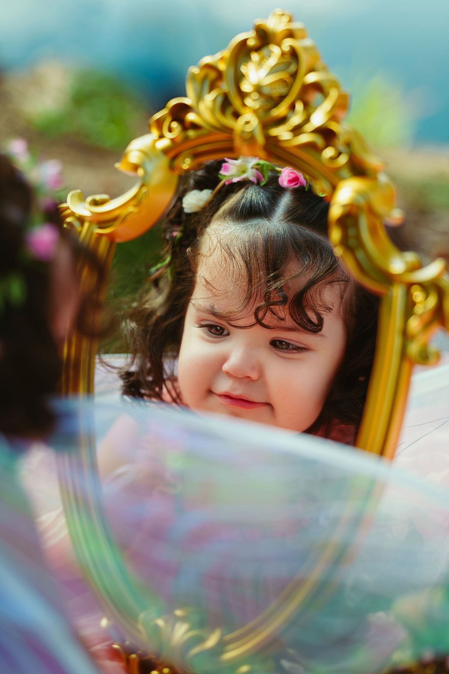 A cute toddler girl with flowers in her hair smiles at her reflection in an ornate gold mirror outdoors.
