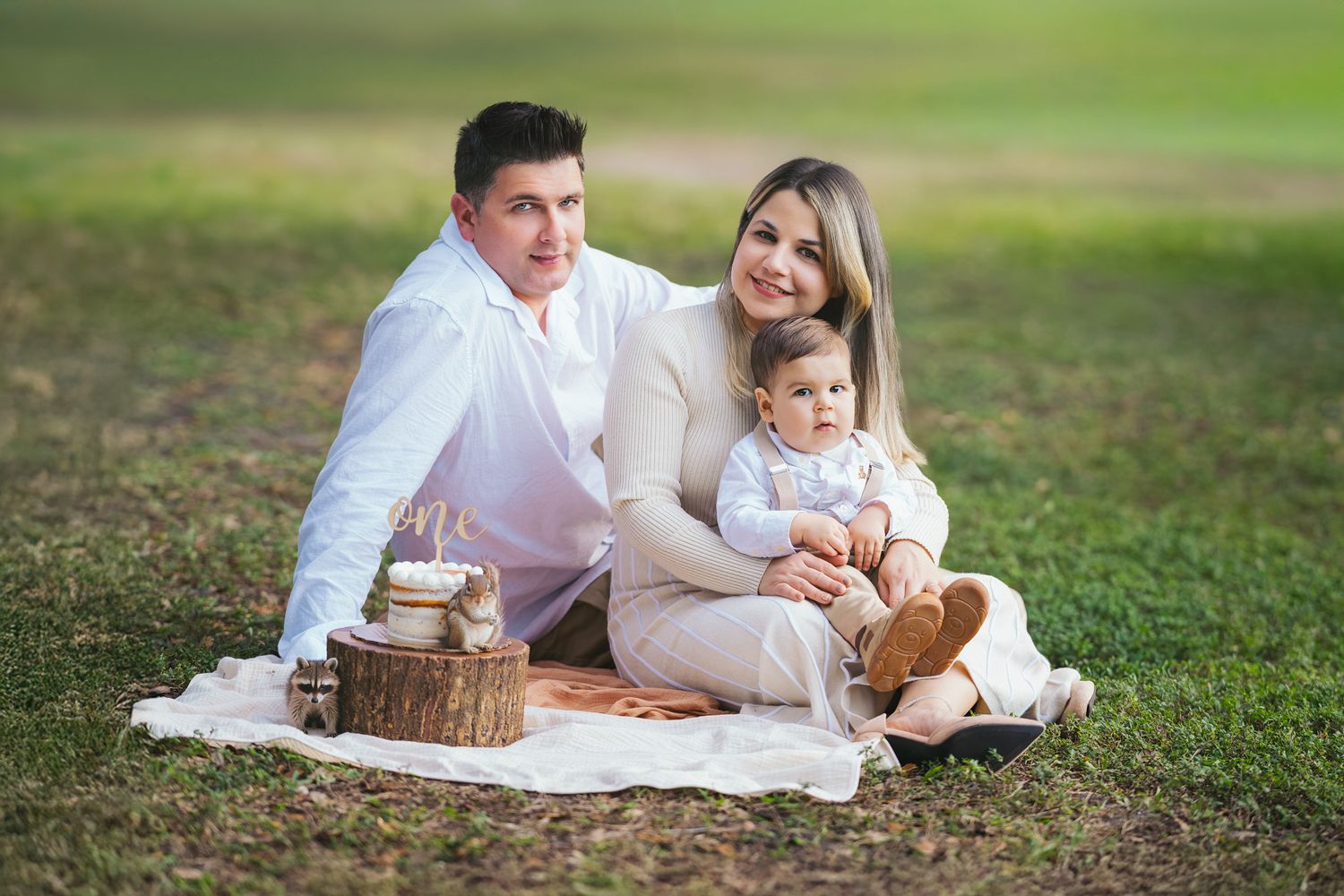 A family wearing white outfits enjoys a picnic on a blanket in a grassy field on a sunny day.