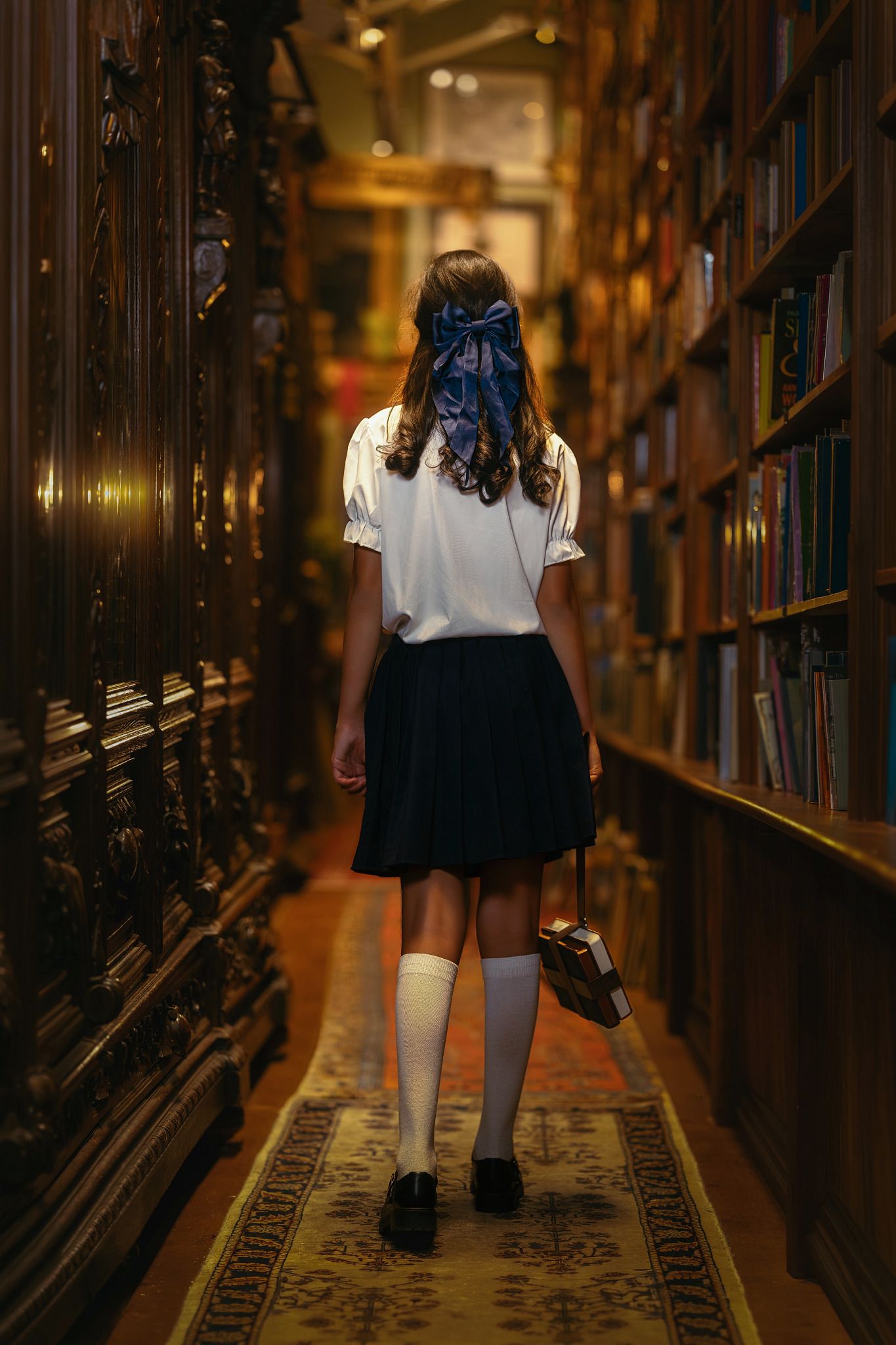 A schoolgirl in white blouse and navy skirt walks through an ornate wooden library holding a book.