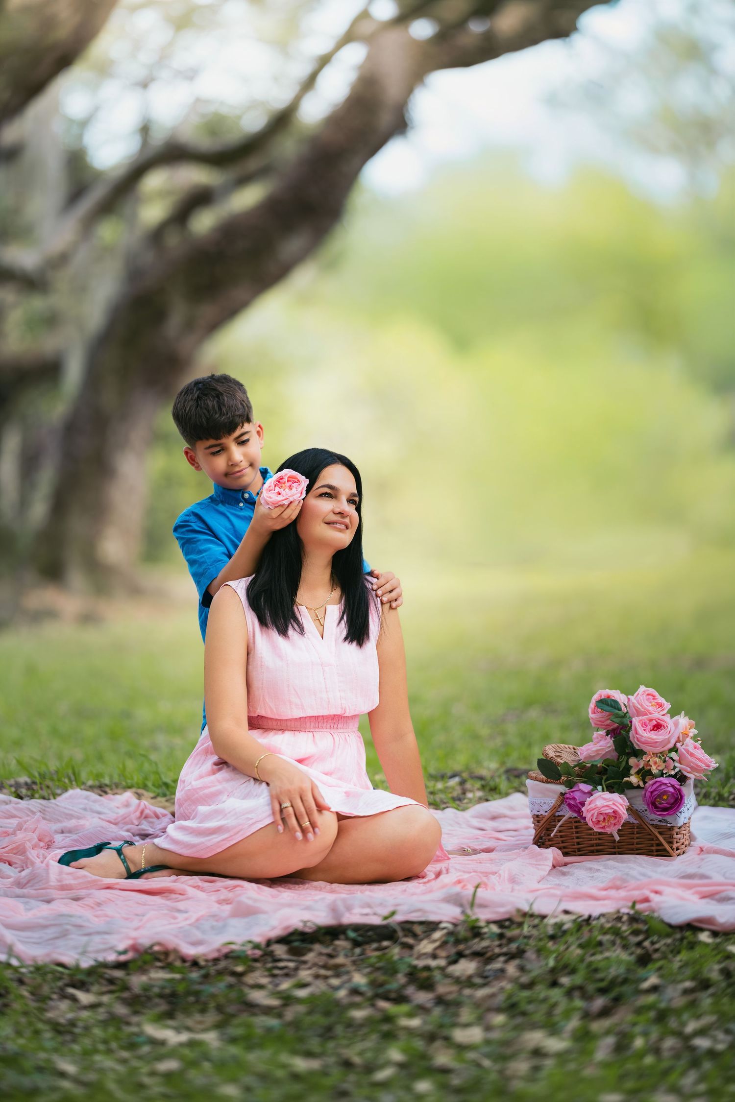 A boy places a pink flower in his mother's hair during a picnic on a pink blanket in a lush green park.