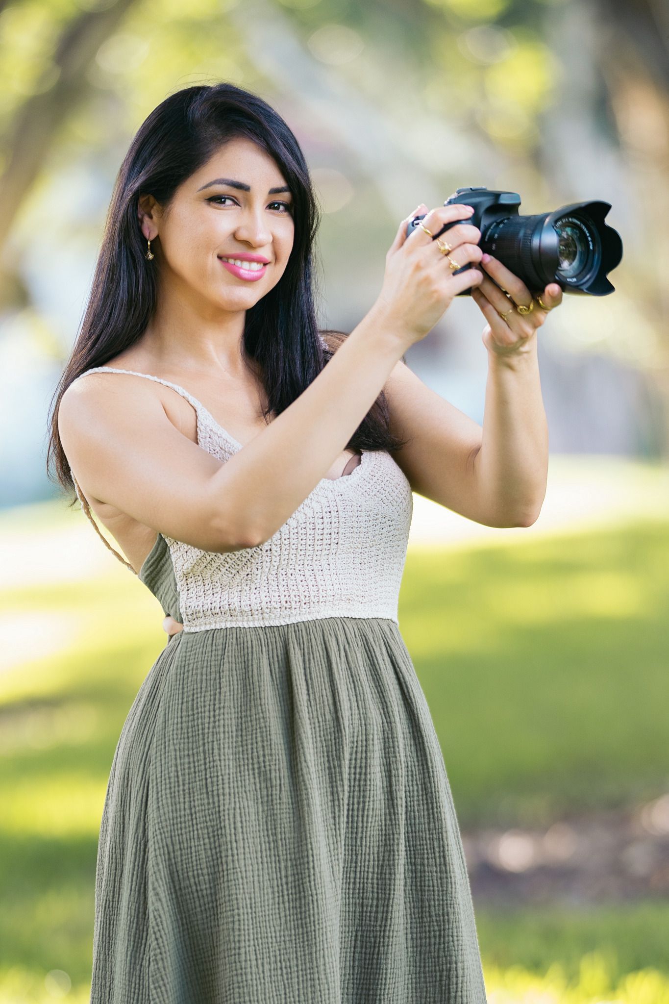 Photographer in olive green dress holds professional DSLR camera while standing in sunny outdoor setting.