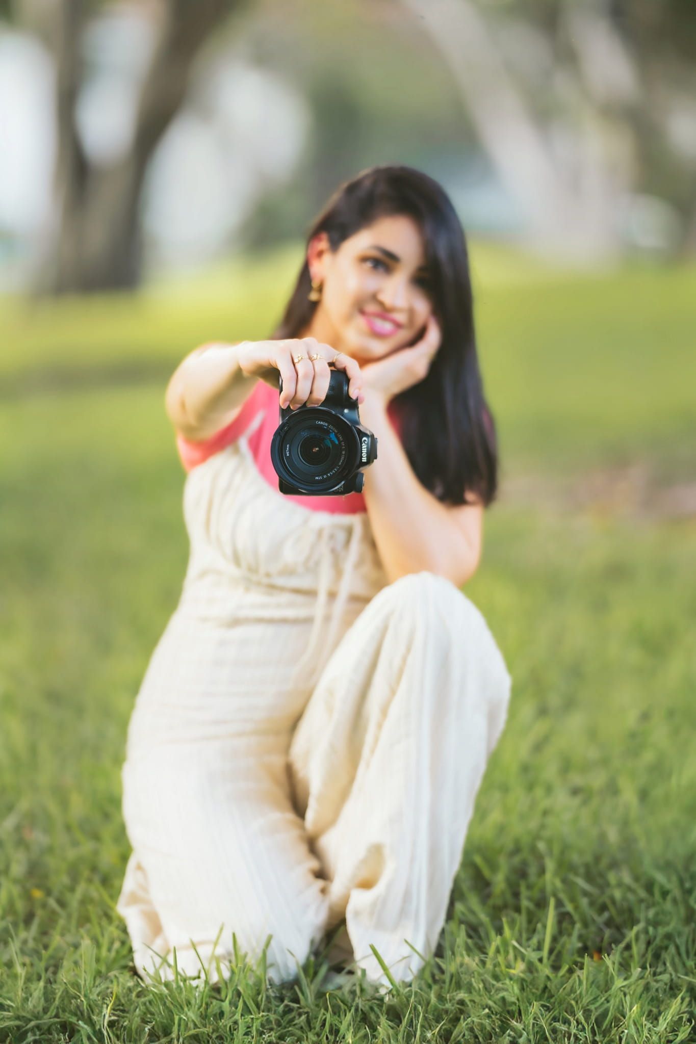 Woman in cream dress sitting on the grass while holding professional camera.