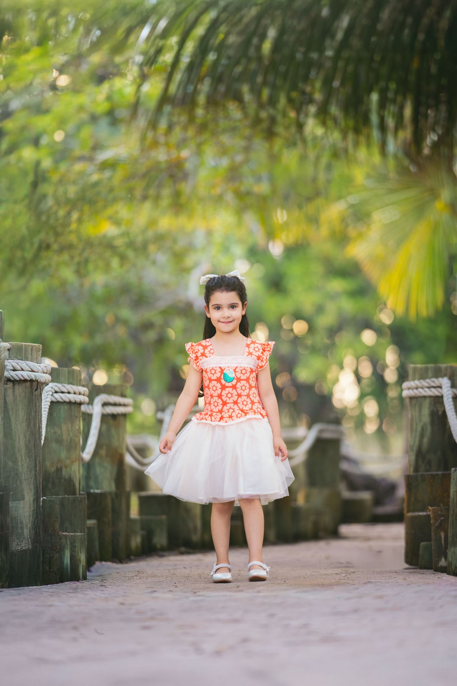 Smiling young girl in orange floral top and white tutu skirt walking along a tropical garden path.