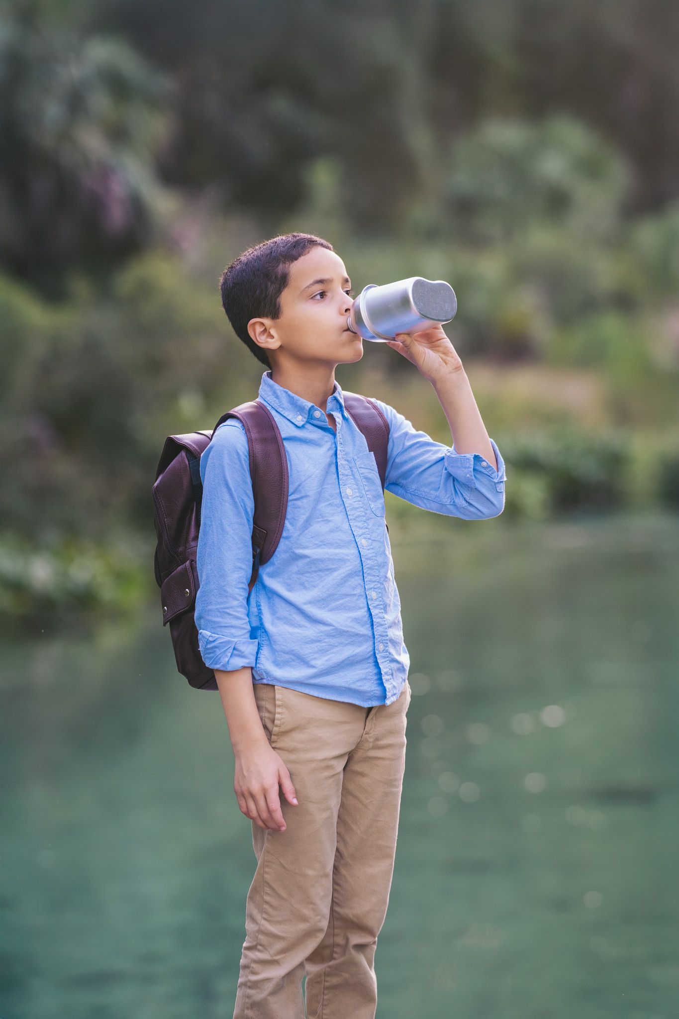 A young boy with a backpack drinks from a metal water bottle outdoors near a scenic green lake.