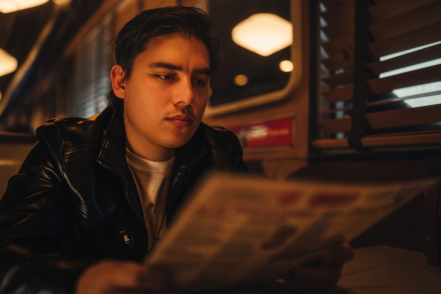 Young man in black leather jacket reading a newspaper in a warmly lit retro diner booth.