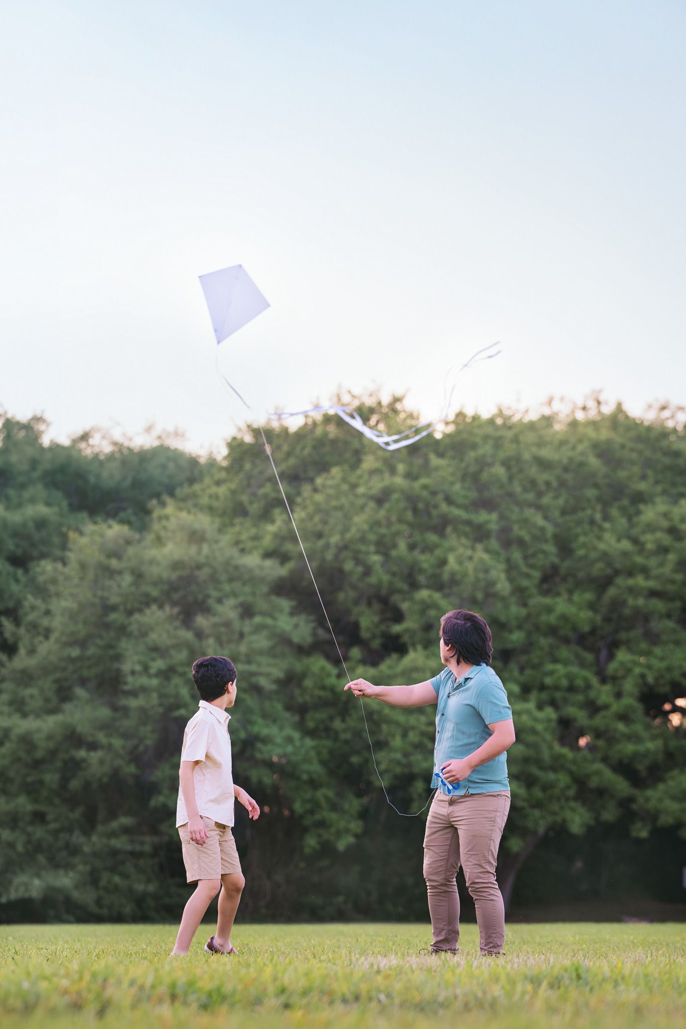 Father and son flying a white kite together in a green park on a clear day.