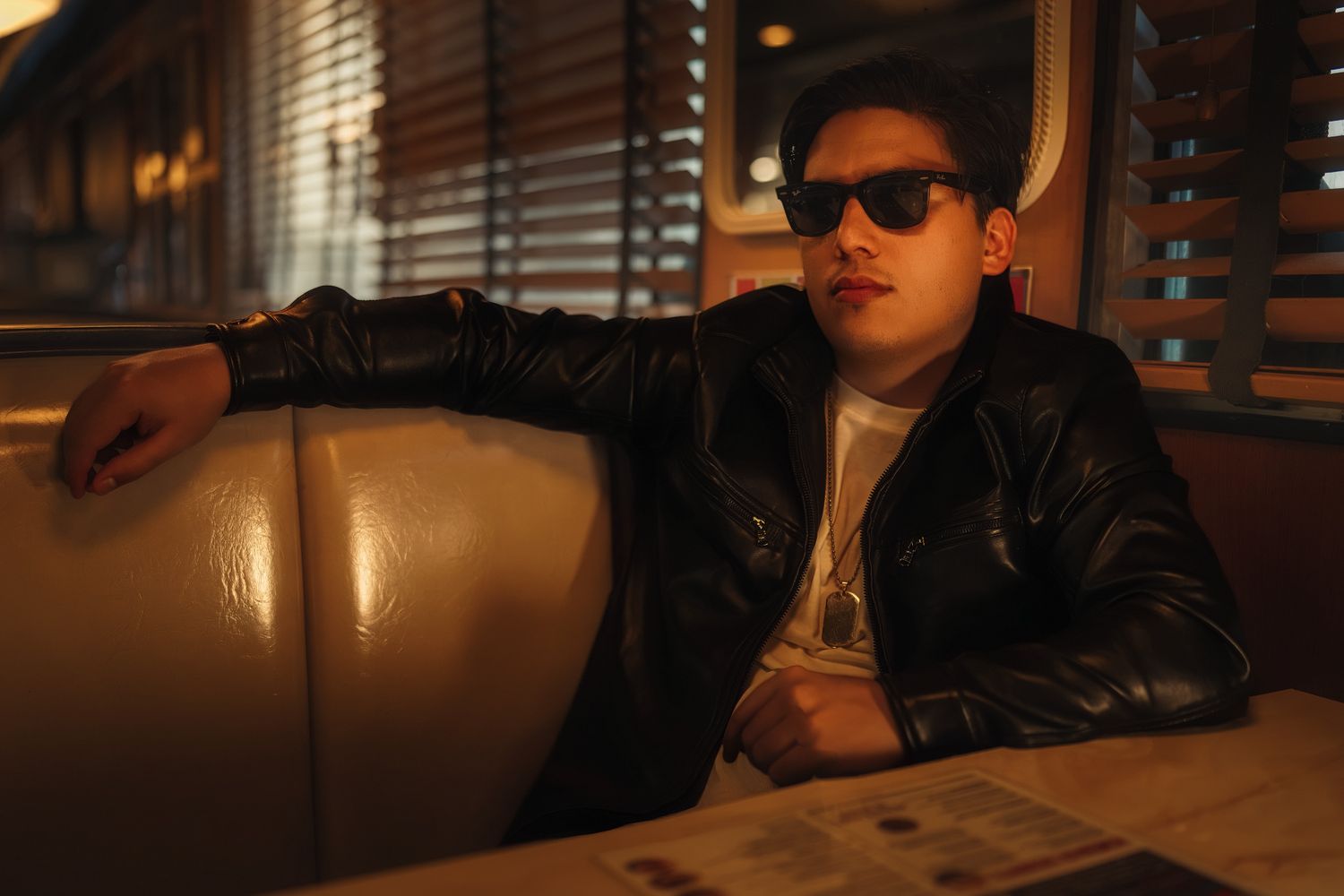 Young man in black leather jacket and sunglasses sitting in a diner booth with warm ambient lighting.