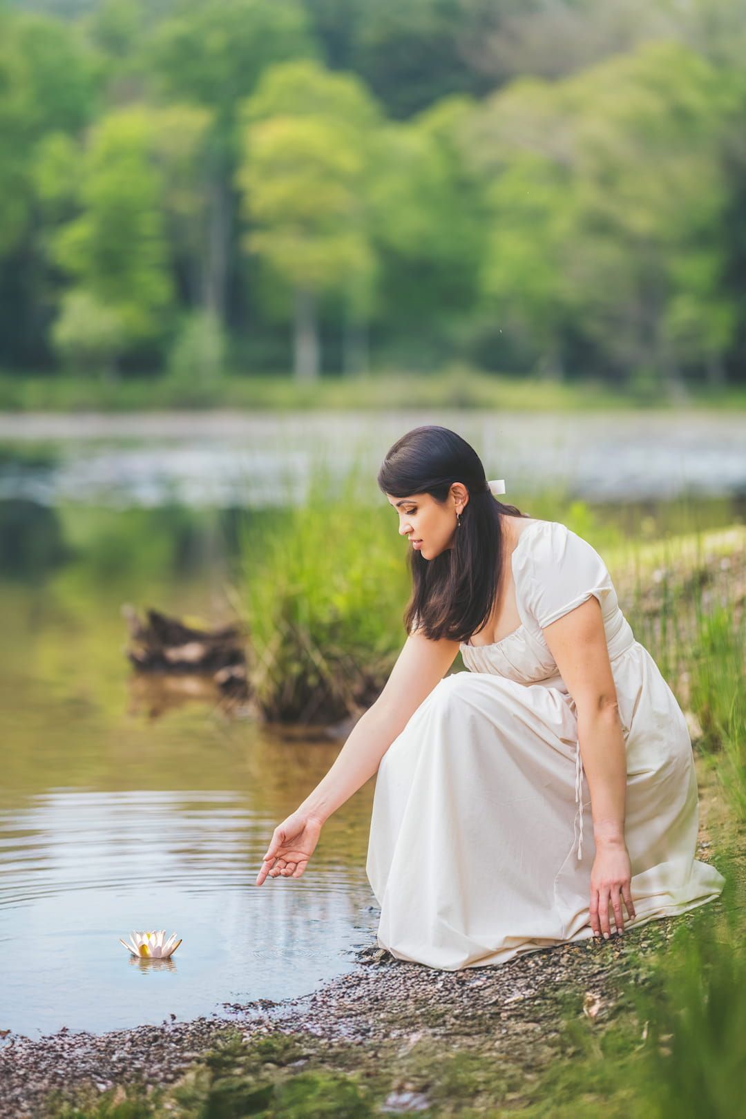 Woman in beige dress kneels by a lakeside, gently touching the water during golden hour.