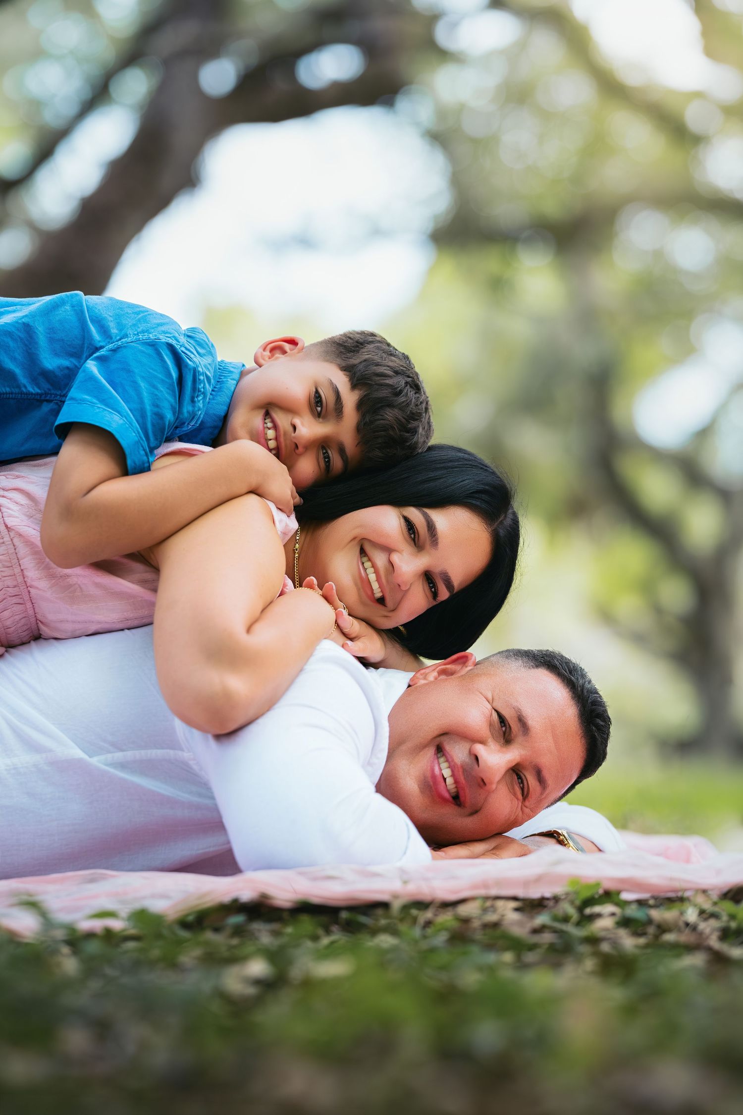 Happy family of three lying in a park, with child on top of smiling parents on a pink blanket outdoors.