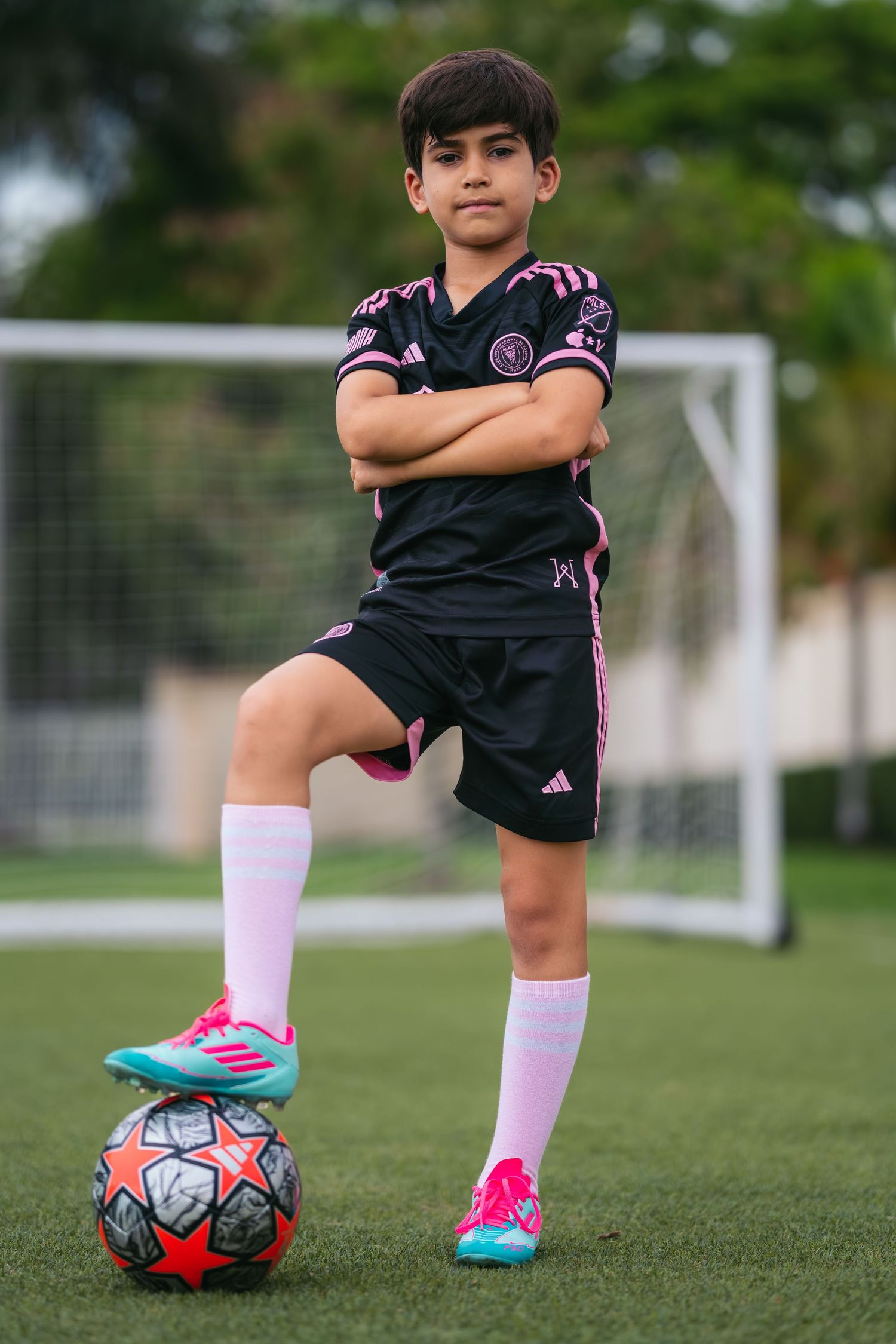 Young soccer player posing with a ball on green grass in front of a goal.