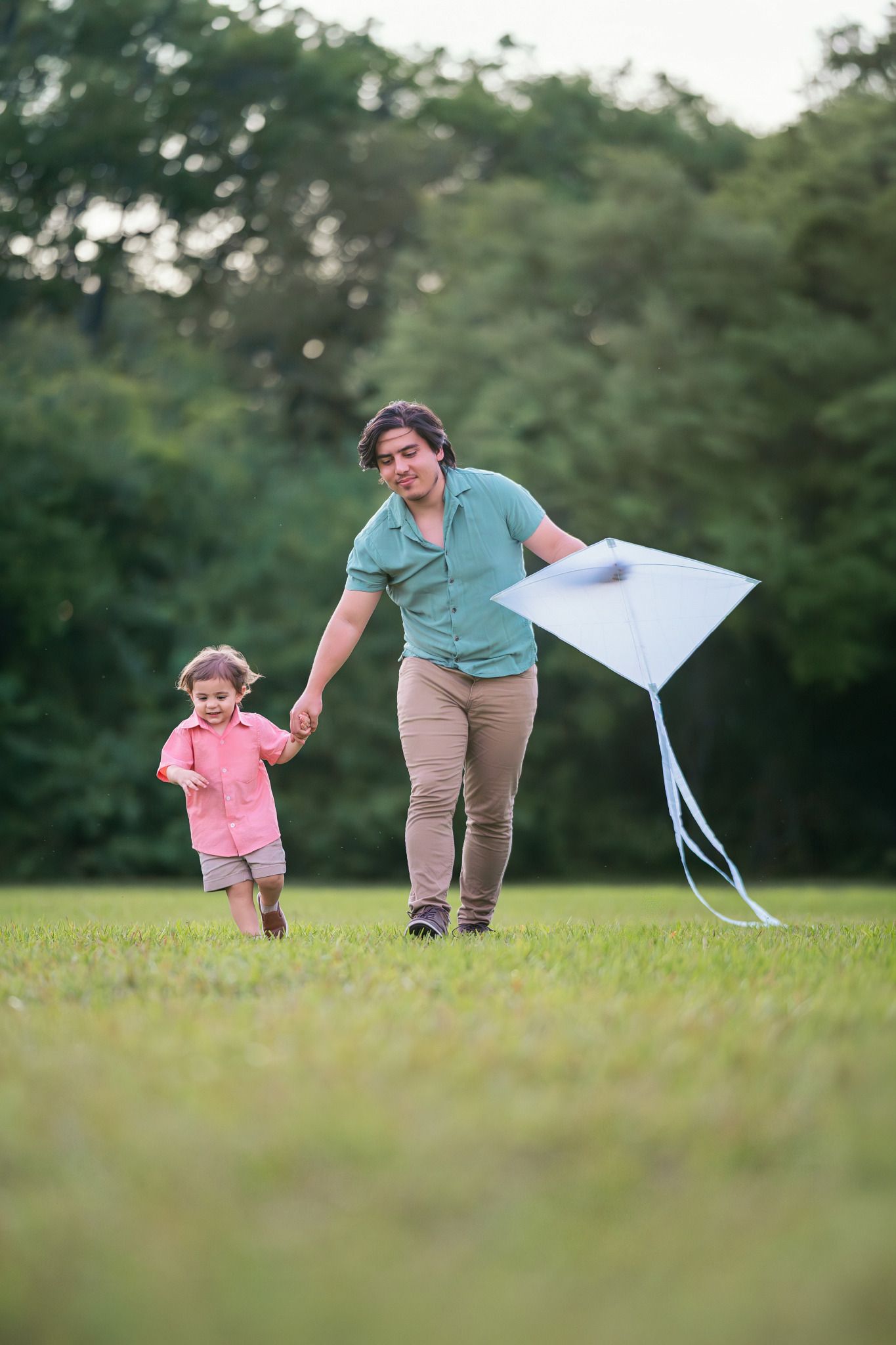 A parent and young child run together on green grass in a park, flying a white kite on a cloudy day.