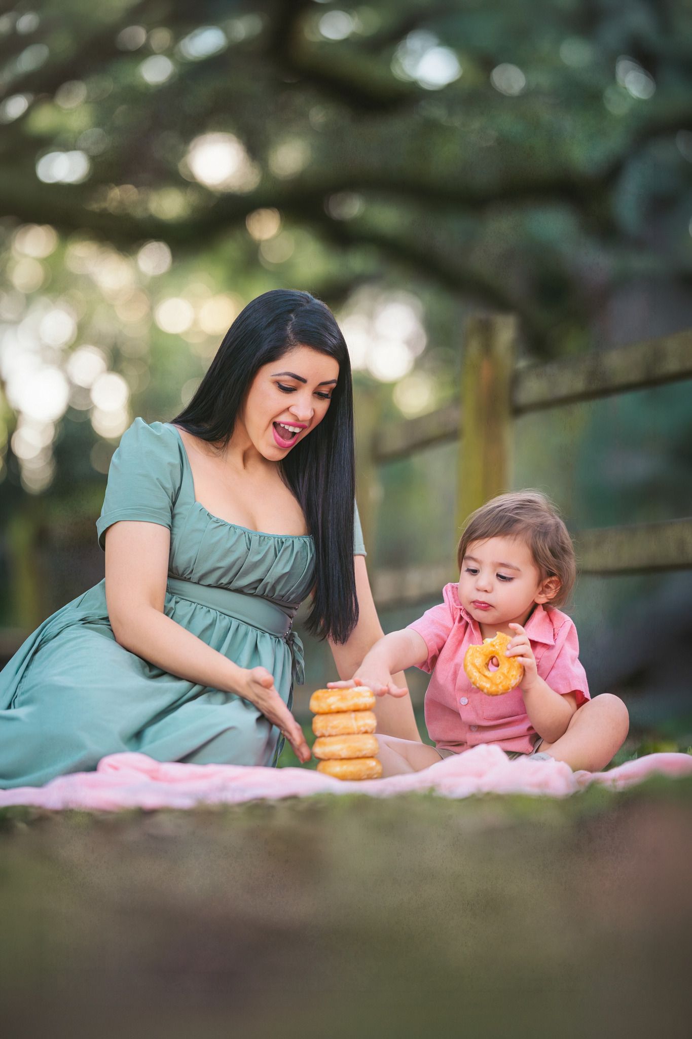 A woman in a green dress sits outdoors with a toddler on a pink blanket, sharing stacked donuts in a park setting.