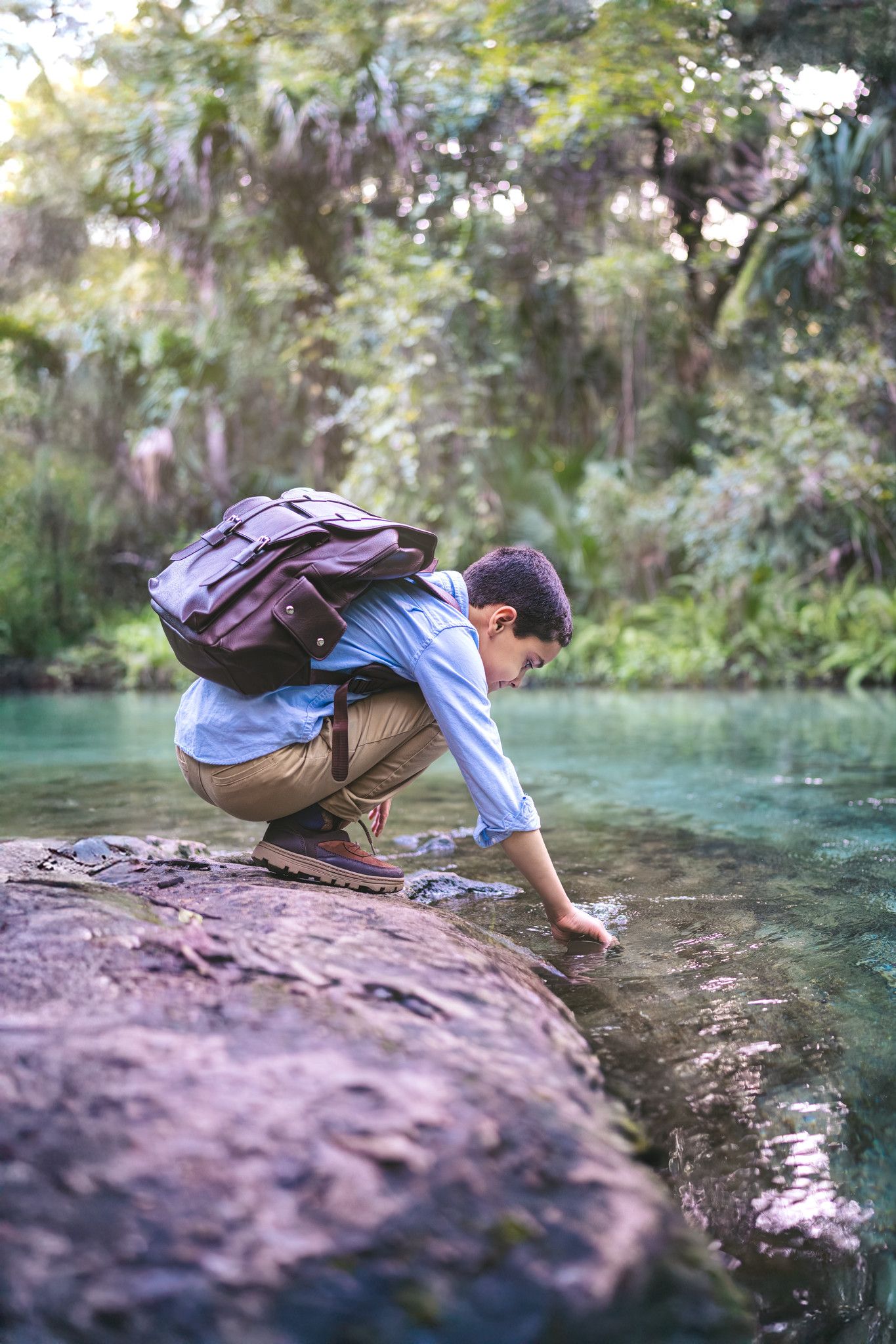 Student with backpack crouches at the edge of a turquoise stream in a lush forest setting.