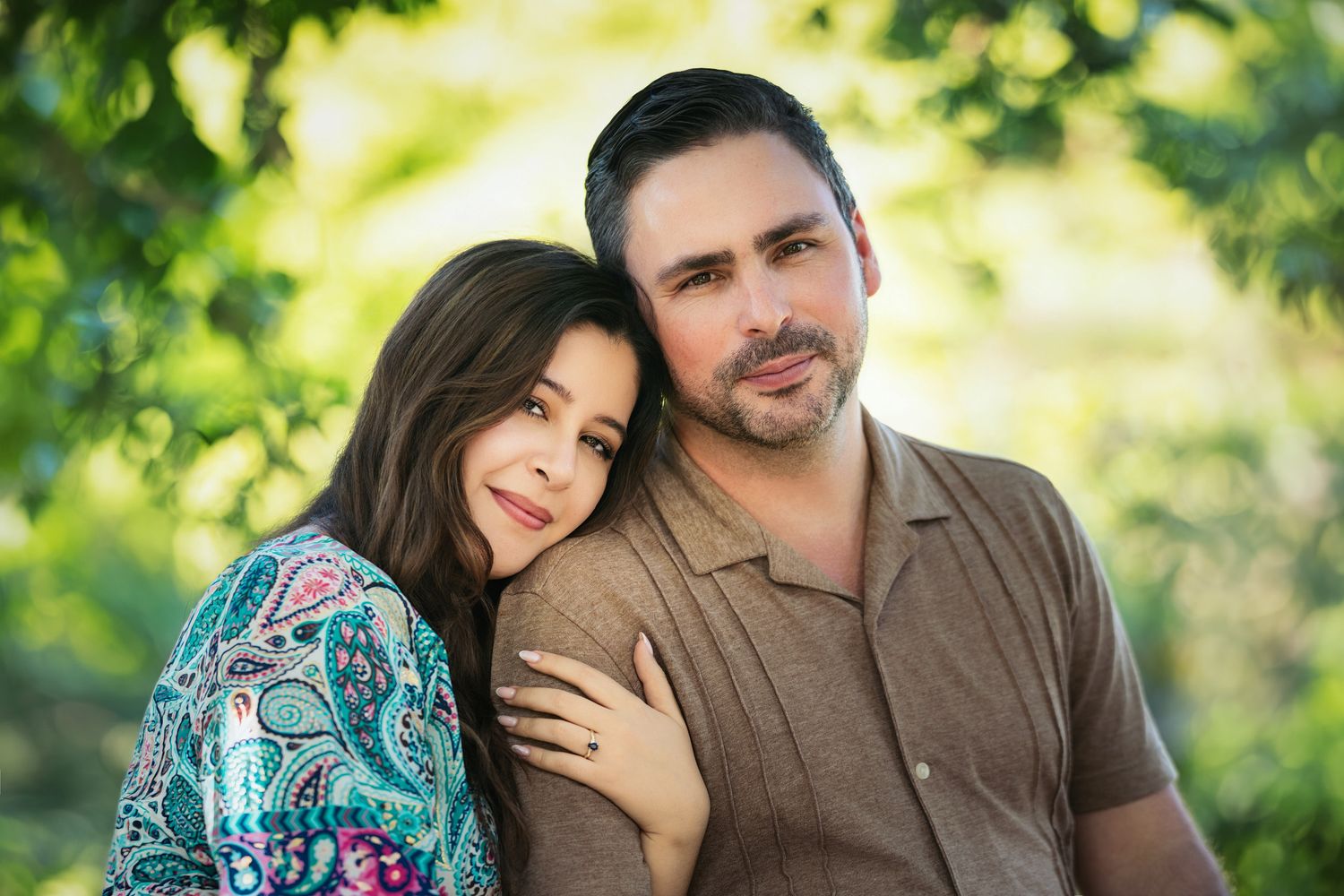 A woman leans on a man's shoulder as they pose together outdoors surrounded by lush green trees.
