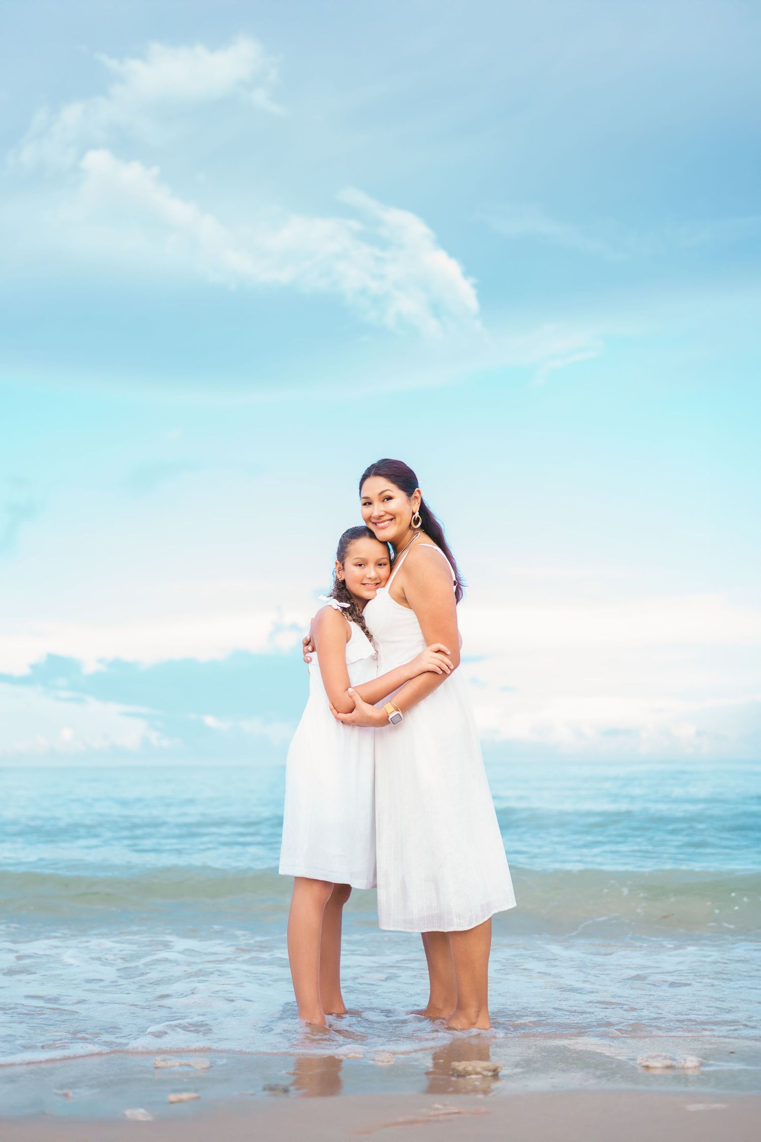 Two people in white dresses embrace while standing in shallow ocean water.